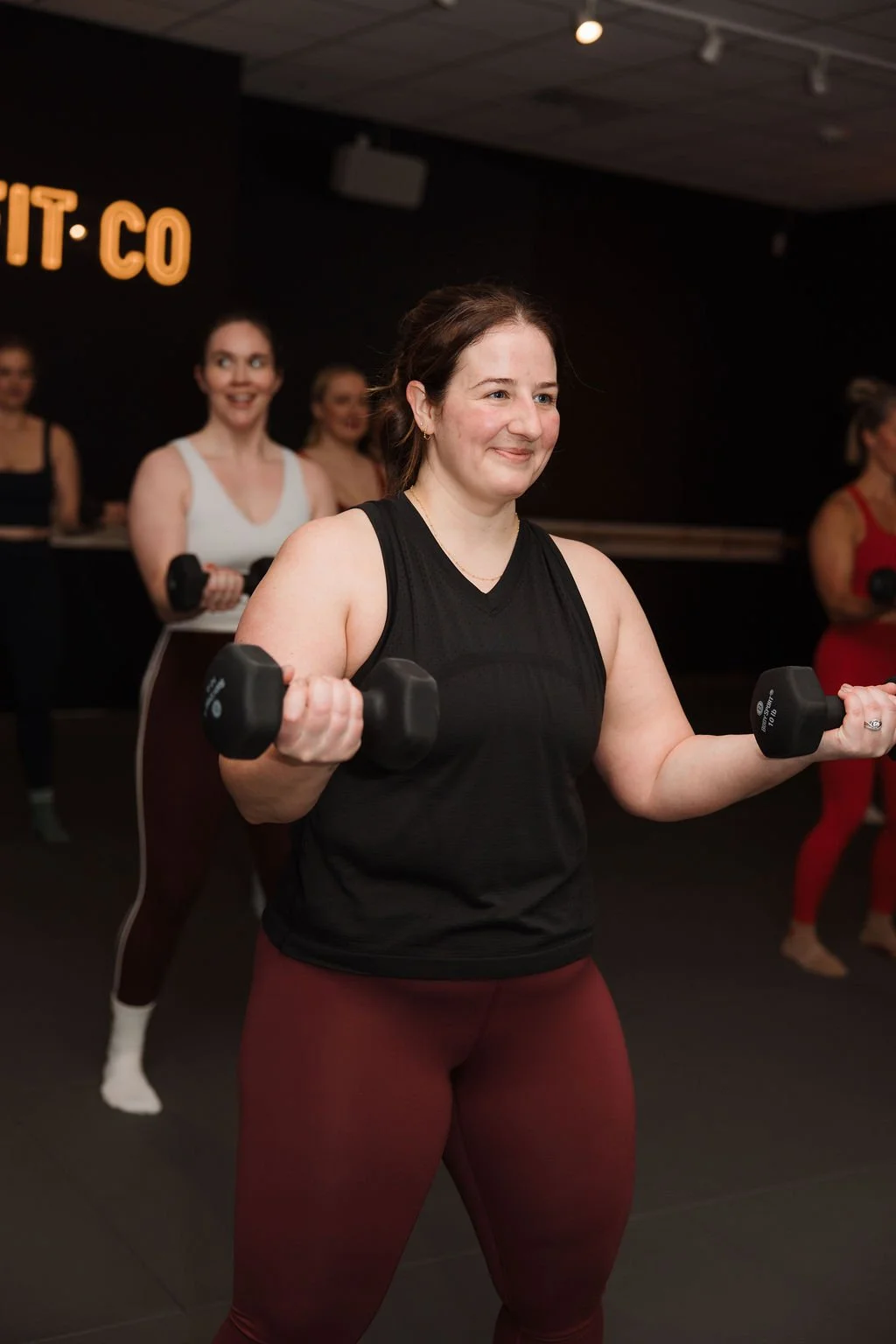 Women participating in a fitness class, lifting dumbbells, with a sign that reads 'FIT CO' in the background.