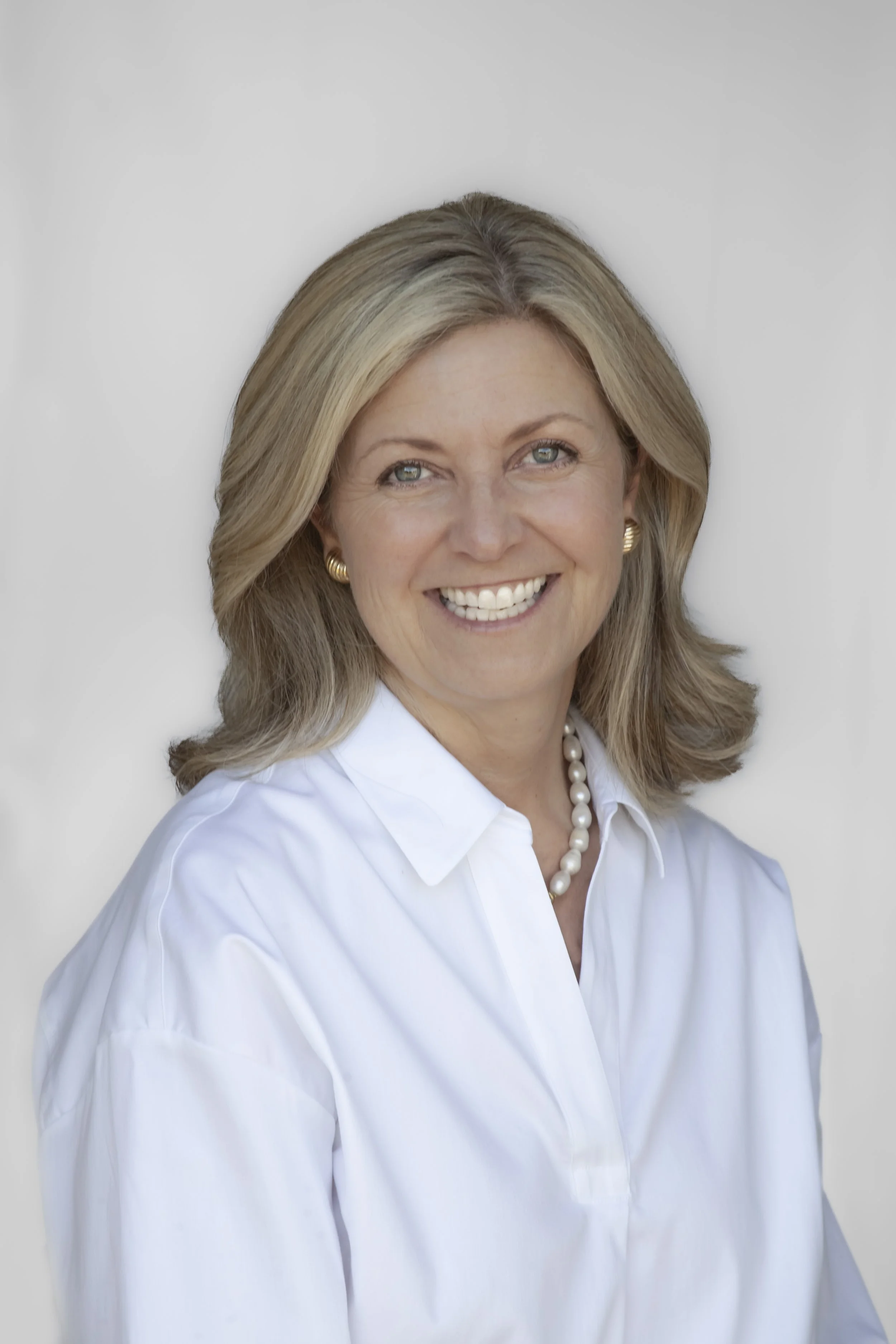 A professional woman with shoulder-length blonde hair, smiling, wearing a dark blue blazer, earrings, and a necklace, against a plain white background.