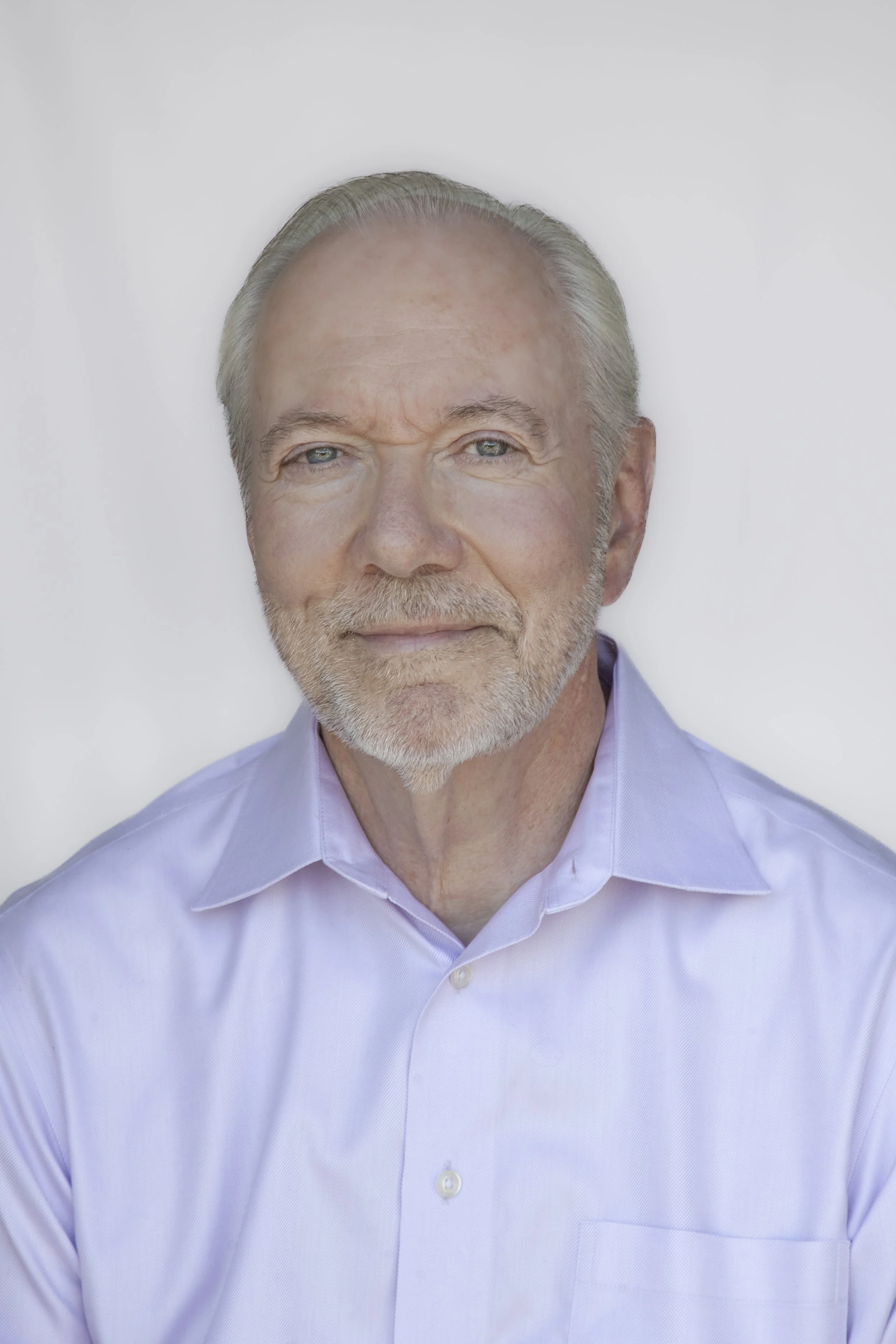 A professional headshot of a man with a beard and short gray hair, wearing a dark blazer and a light-colored shirt, against a plain light blue background.