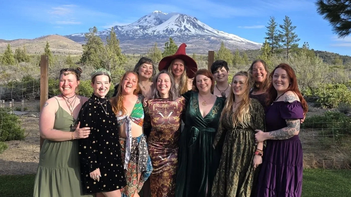 A group of ten women, dressed in colorful and diverse outfits, standing outdoors in front of Mount St. Helens and a clear blue sky, smiling and posing for a photo.