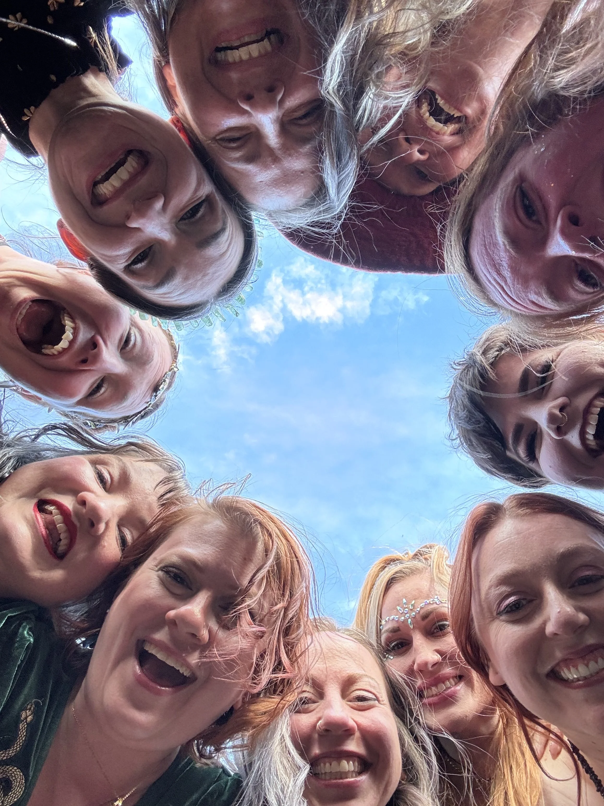 Group of smiling women with colorful hair, some wearing accessories, looking down at the camera, with a blue sky in the background.
