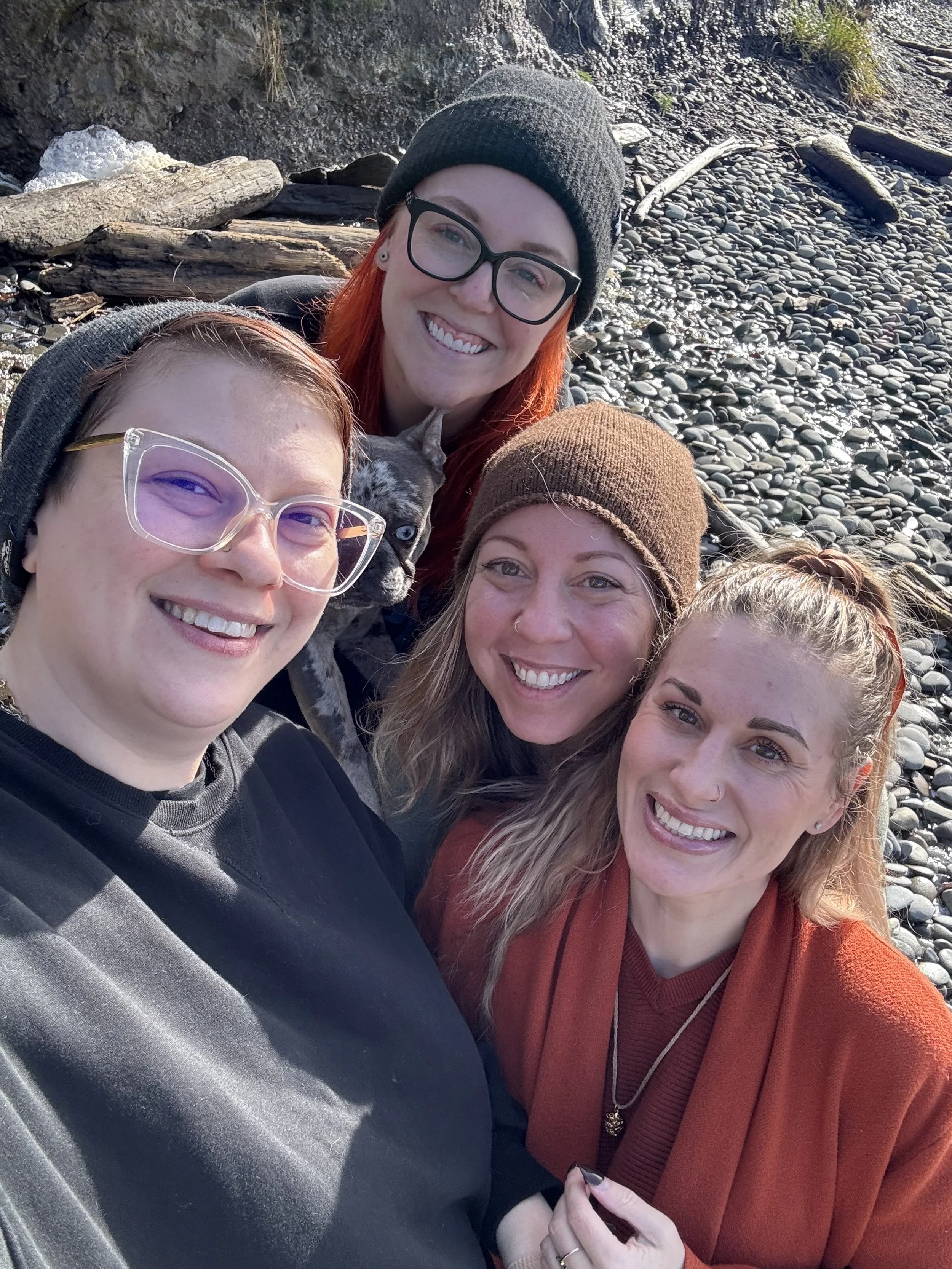 Four women smiling for a selfie on a pebble beach with driftwood and a small dog in the background.