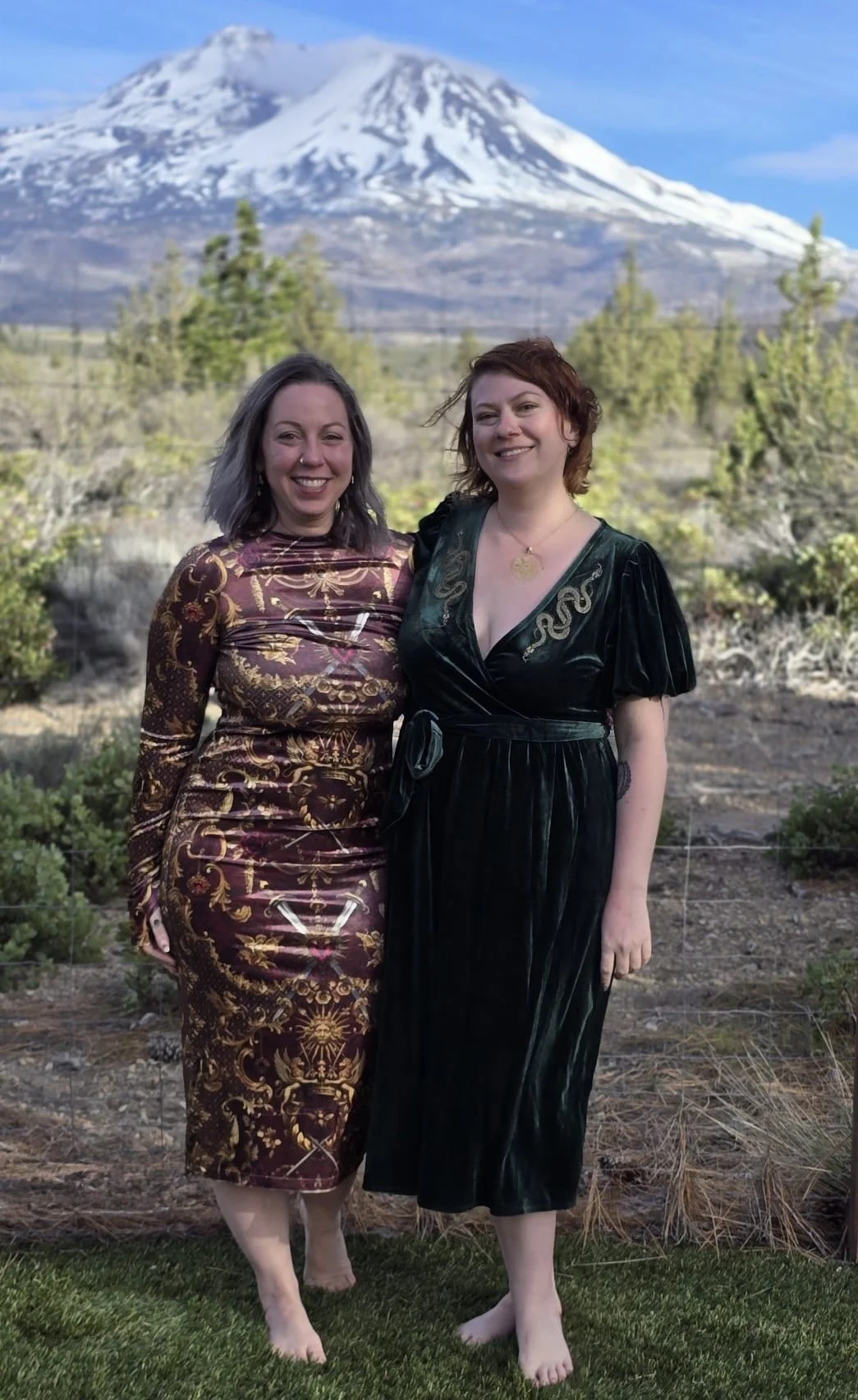 Two women standing outdoors in front of Mount St. Helens, smiling, with trees and shrubs around them, both barefoot, wearing elegant dresses with intricate patterns.