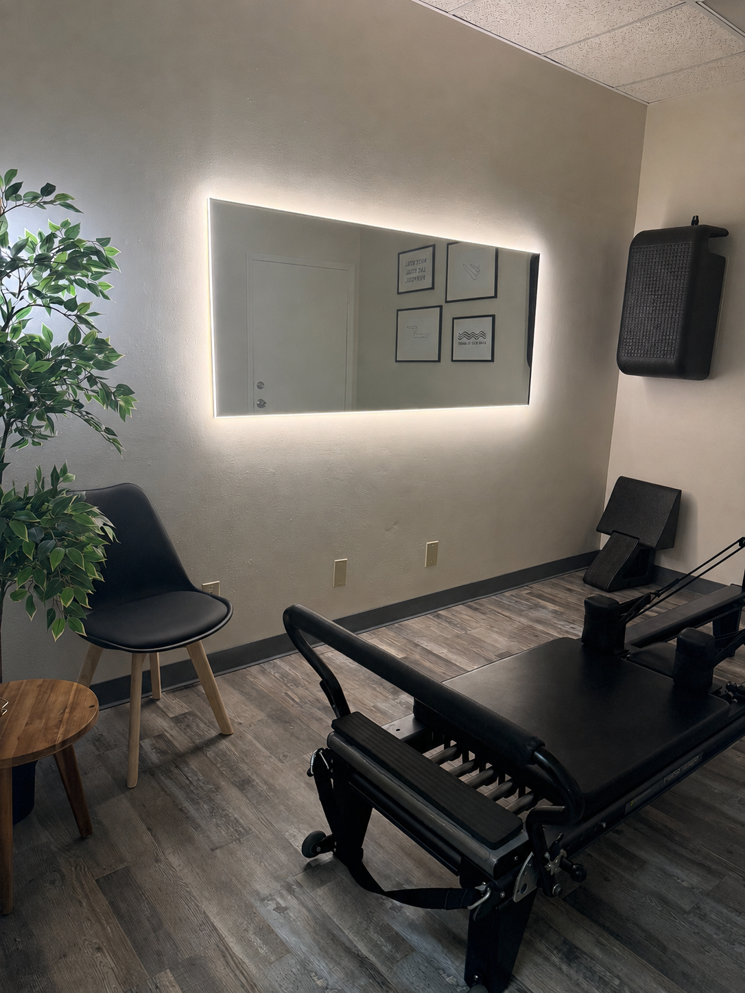 Private pilates studio with a reformer, black chair, small wooden table, large mirror with LED lighting, framed artwork on the wall, potted plant, and mounted black speaker.