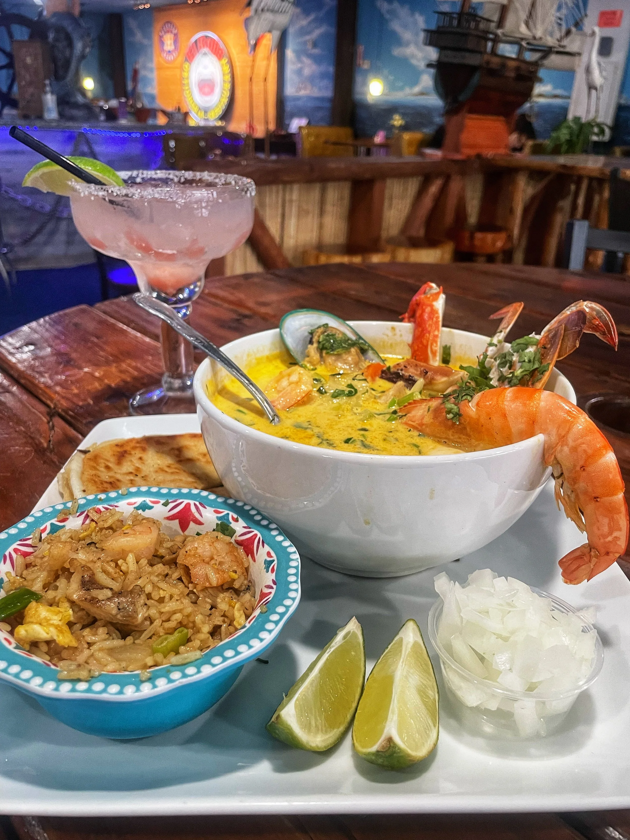 A seafood dish with a large bowl of yellow curry containing shrimp, mussels, and lobster, garnished with cilantro, with lime wedges, onion, and a small bowl of rice and flatbread on a white tray. A pink cocktail with a lime wedge and a straw is on the dark wooden table in a restaurant setting.
