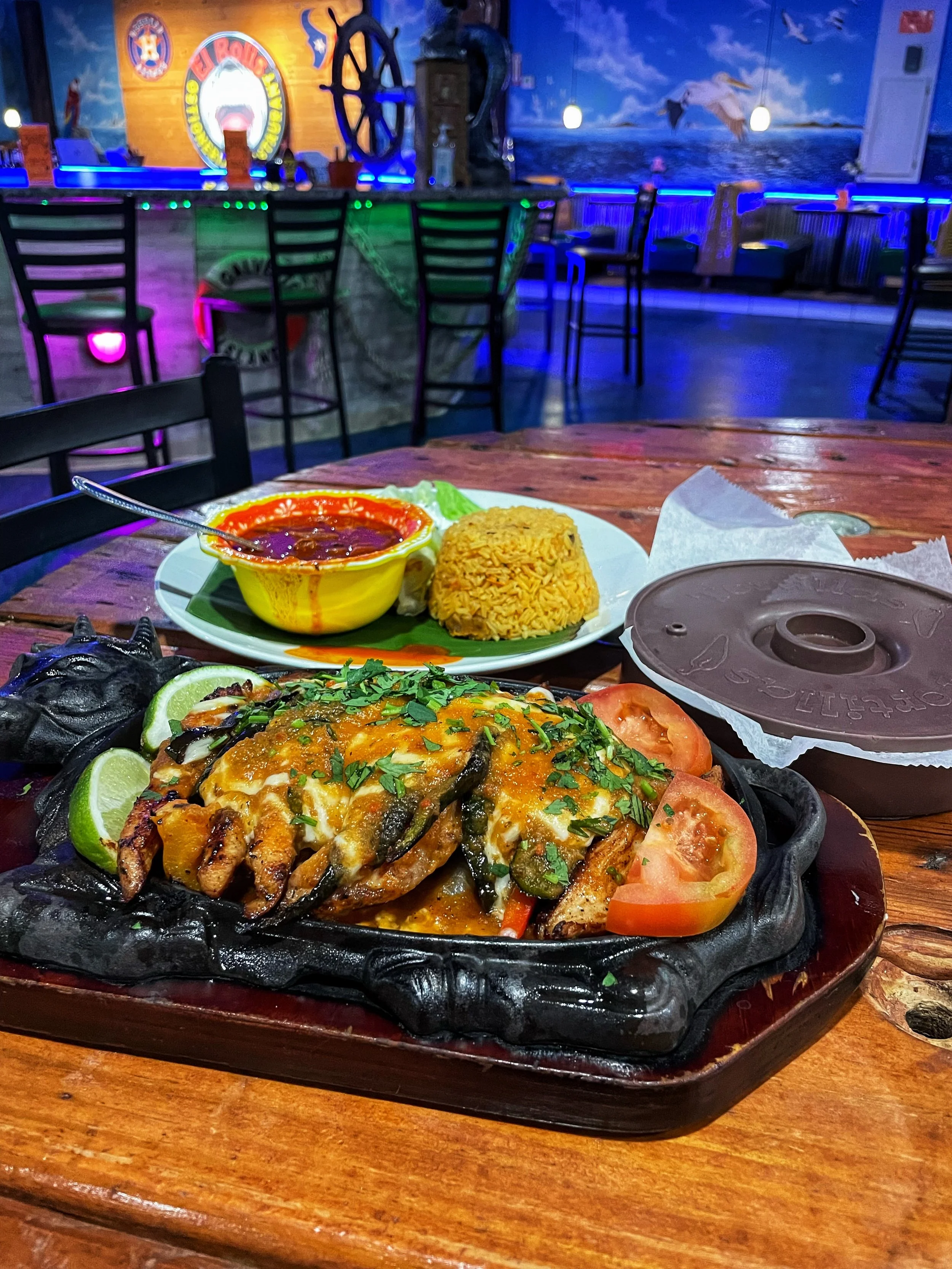 Mexican seafood dish on a sizzling platter with tomatoes, lime, and cilantro, accompanied by Mexican rice, a bowl of red sauce, and a chocolate flan dessert on a table in a colorful restaurant.