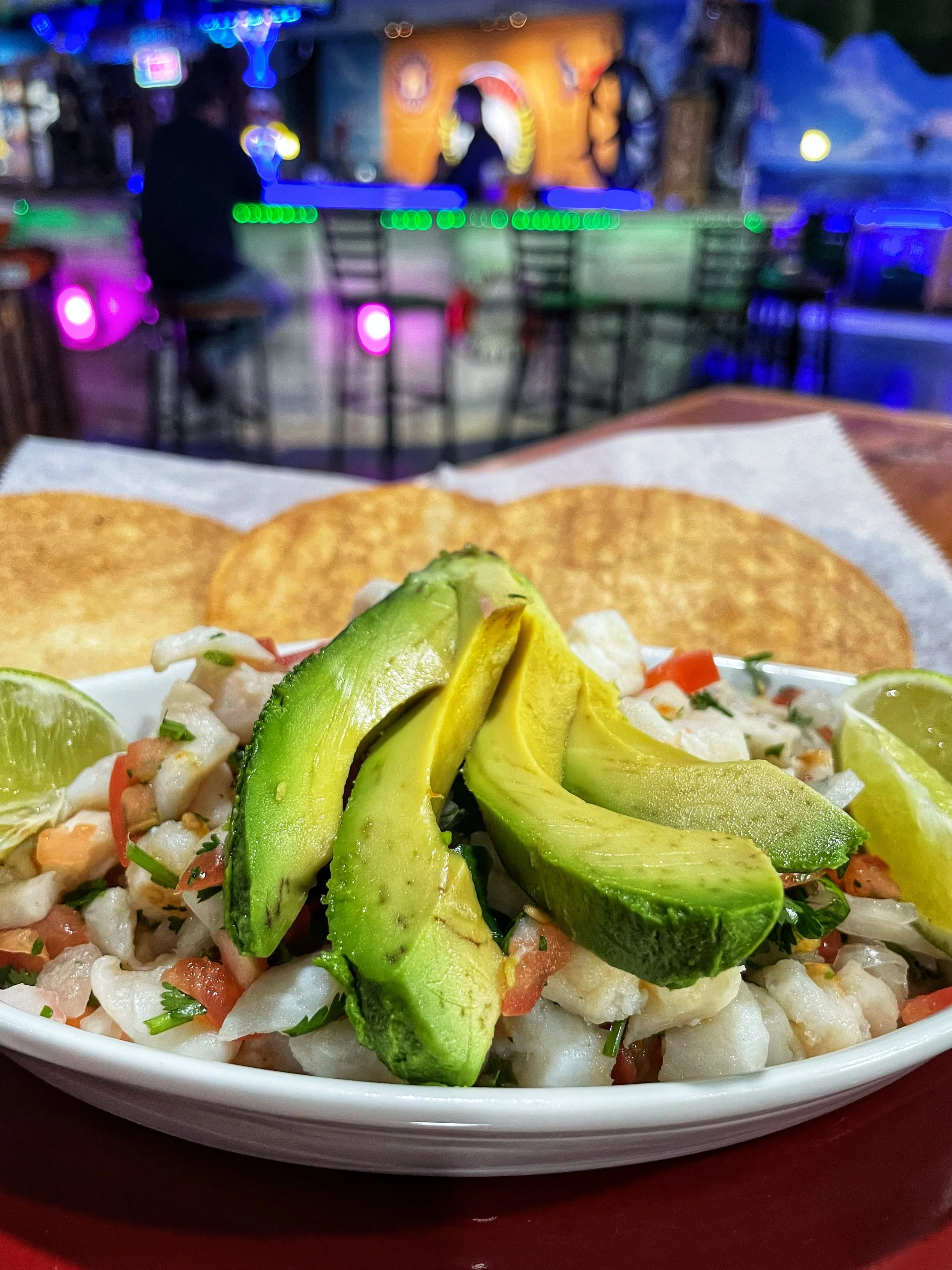 Close-up of a bowl of ceviche garnished with sliced avocado, lime wedges, and tortilla chips with a bar and neon lights in the background.