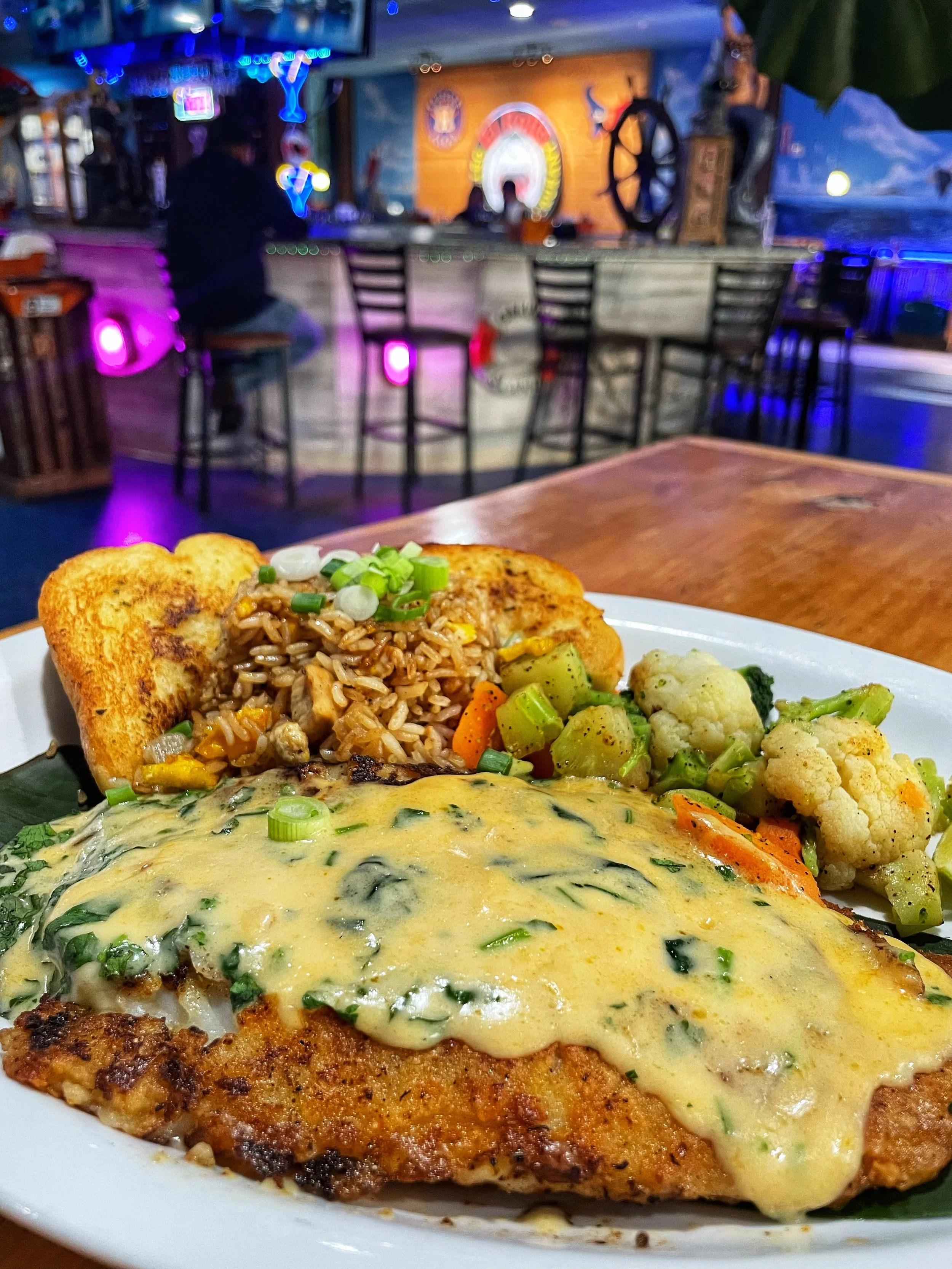 Plate of food featuring a breaded chicken breast topped with creamy spinach sauce, alongside rice with vegetables, roasted cauliflower, and a piece of toast, in a restaurant with a bar and neon lighting in the background.