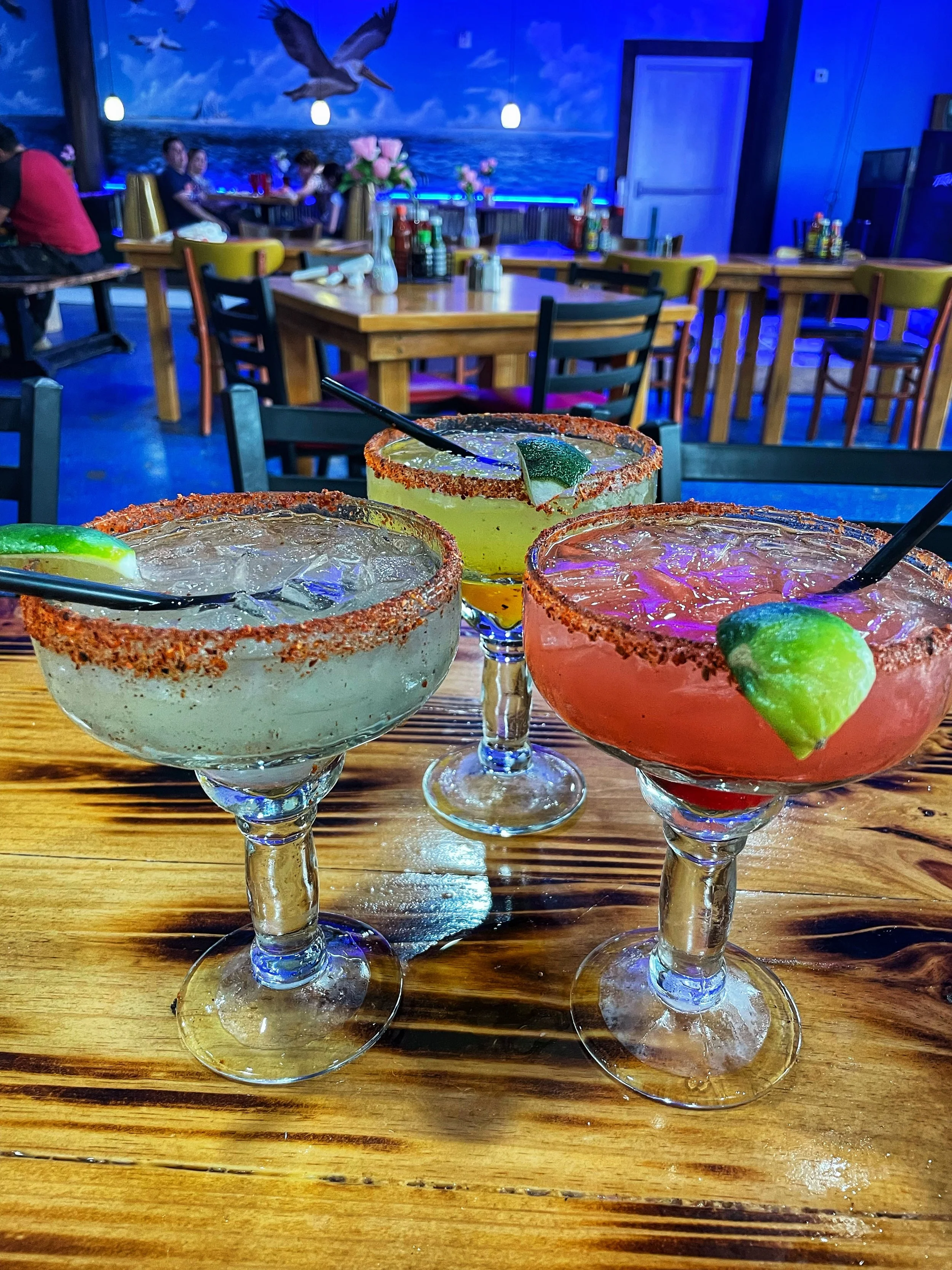 Three colorful margaritas with salted rims and lime garnishes on a wooden bar counter in a restaurant or bar with blue lighting and nautical wall decor.
