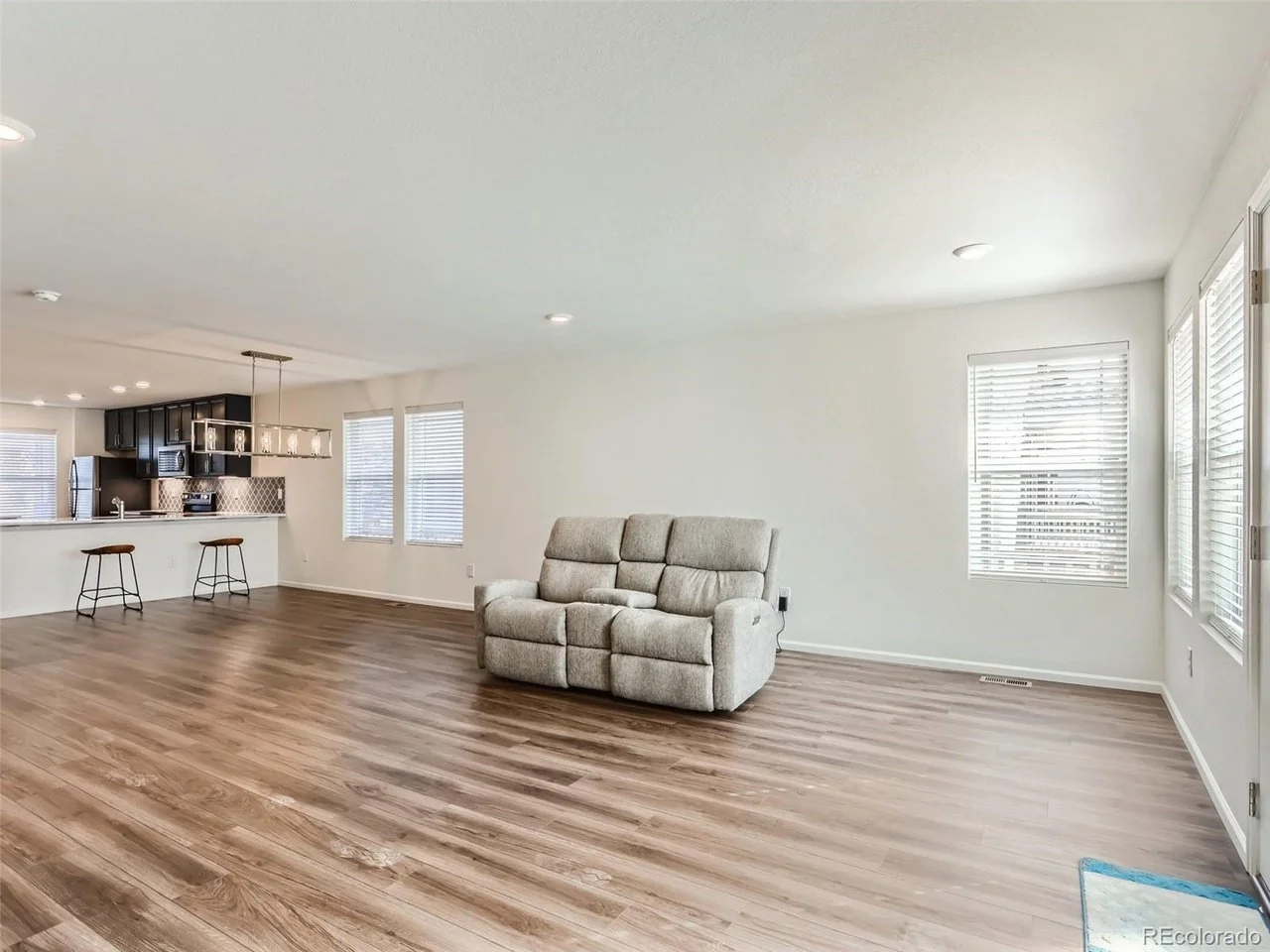 Spacious open living room with light wood flooring, gray recliner sofa, large windows with blinds, and view into a modern kitchen with bar stools and pendant lighting.