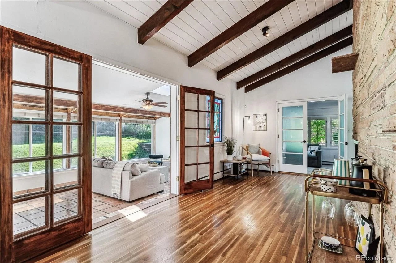 Interior of a living room with wooden floors, exposed beams, and a stone wall. It has glass French doors, a sunroom with sofas and a ceiling fan, and a small sitting area with a chair and side table. There is a bar cart and decorative items along the wall.