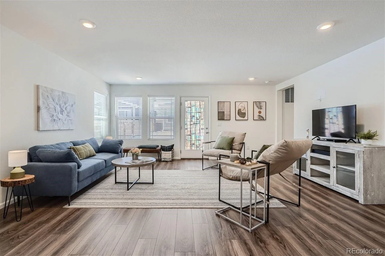 Modern living room with blue sofa, armchairs, round coffee table, TV on white stand, artwork on walls, wooden flooring, and large windows with blinds.