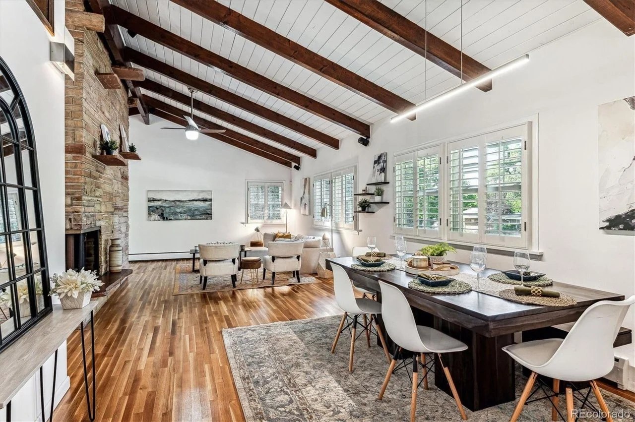 Spacious and modern open-plan living and dining room with wooden floors, vaulted ceiling with exposed beams, stone fireplace, and large windows with white shutters.
