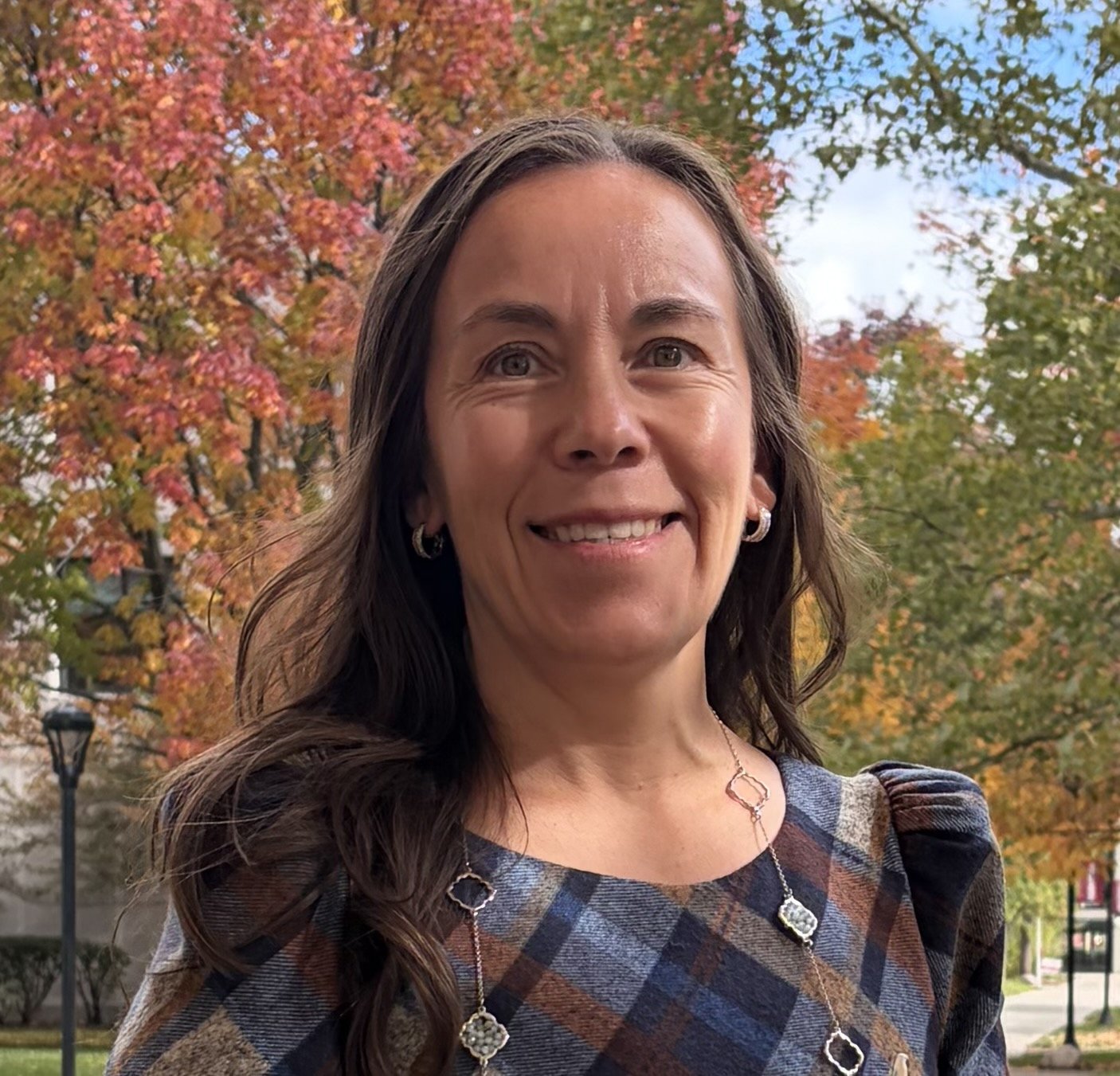 Portrait of a woman with long brown hair, smiling, wearing a green top with sheer sleeves and a silver necklace with two heart pendants, standing outdoors with green foliage in the background. | Beronica Cruz, BGS, IFC