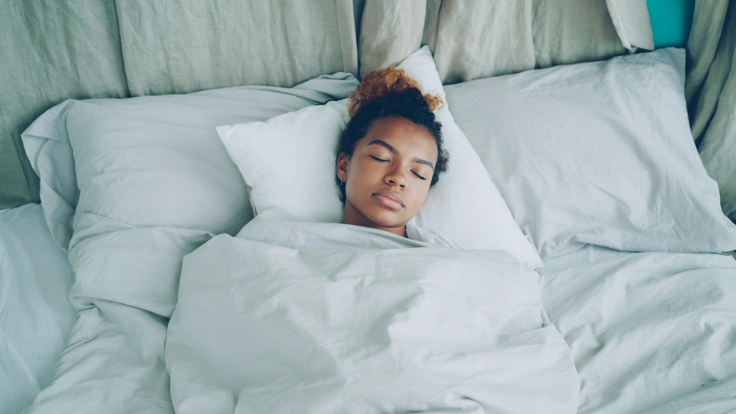 A woman with curly hair and earrings sleeping on a bed with white sheets and pillows.