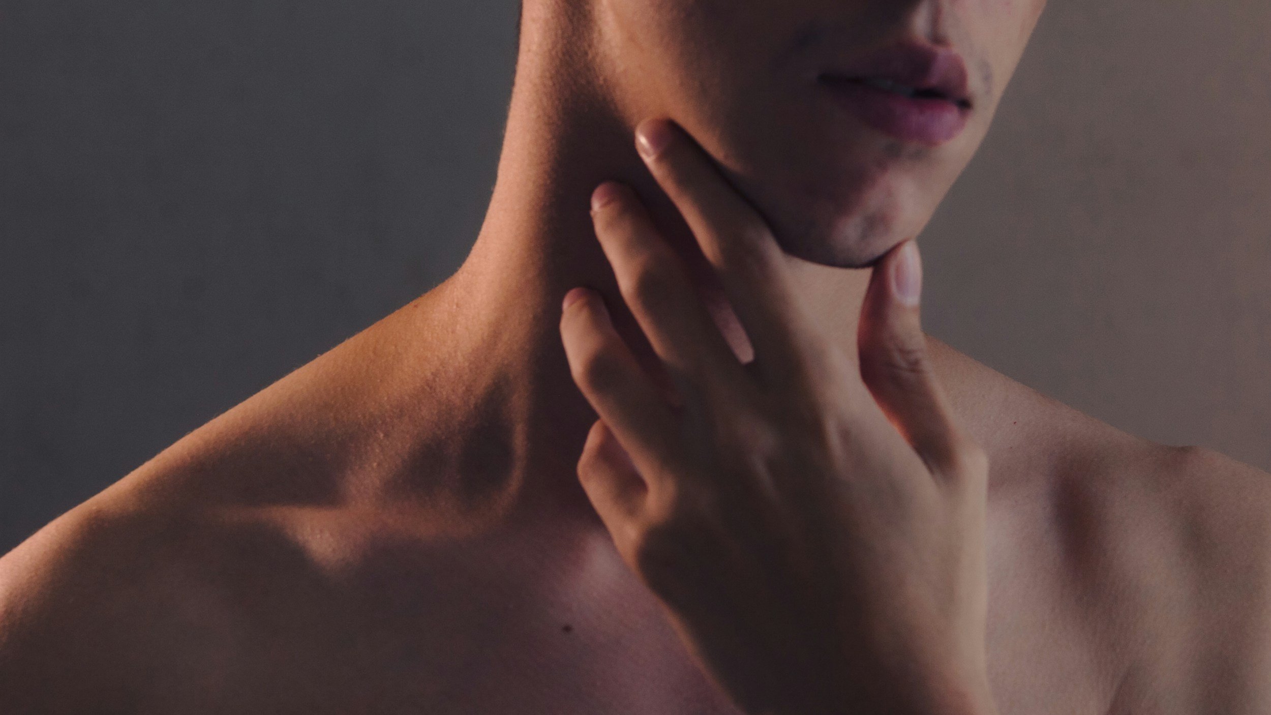 Close-up of a person's face and neck with one hand touching their chin and neck, showing skin and lips, against a dark background.