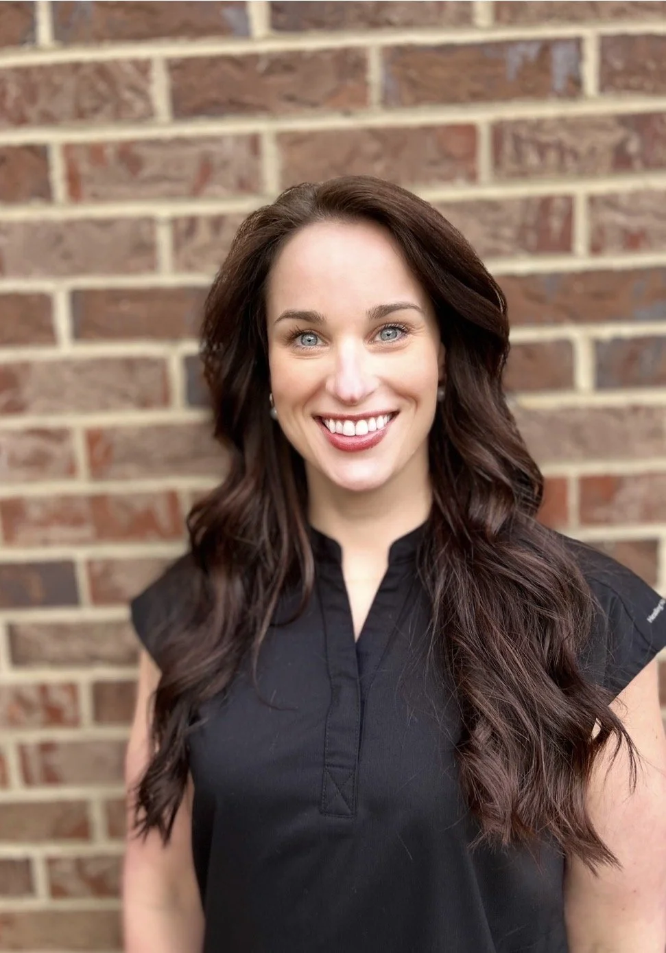 A woman standing in front of a brick wall, smiling, with long, wavy brown hair and wearing a black top.