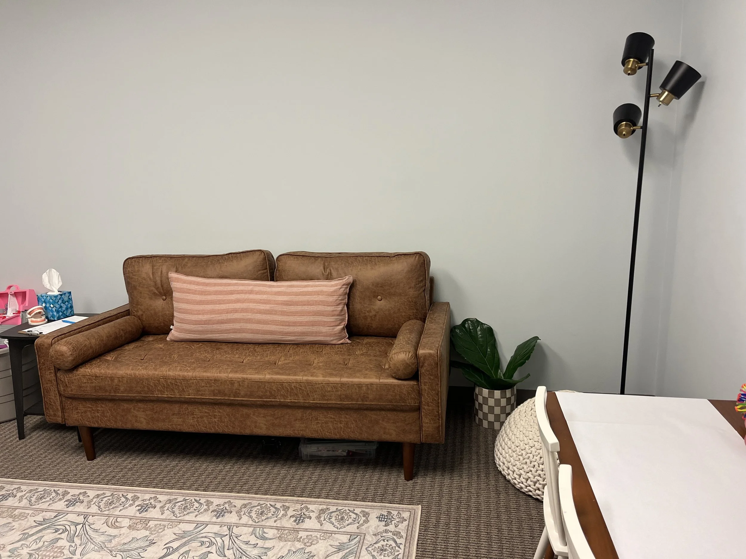 Living room with a brown vintage sofa, a pink striped pillow, a black side table with tissues and toys, a checkered plant pot with a large green plant, a modern black and gold floor lamp, a beige knitted pouf, and part of a dining table with white chairs.