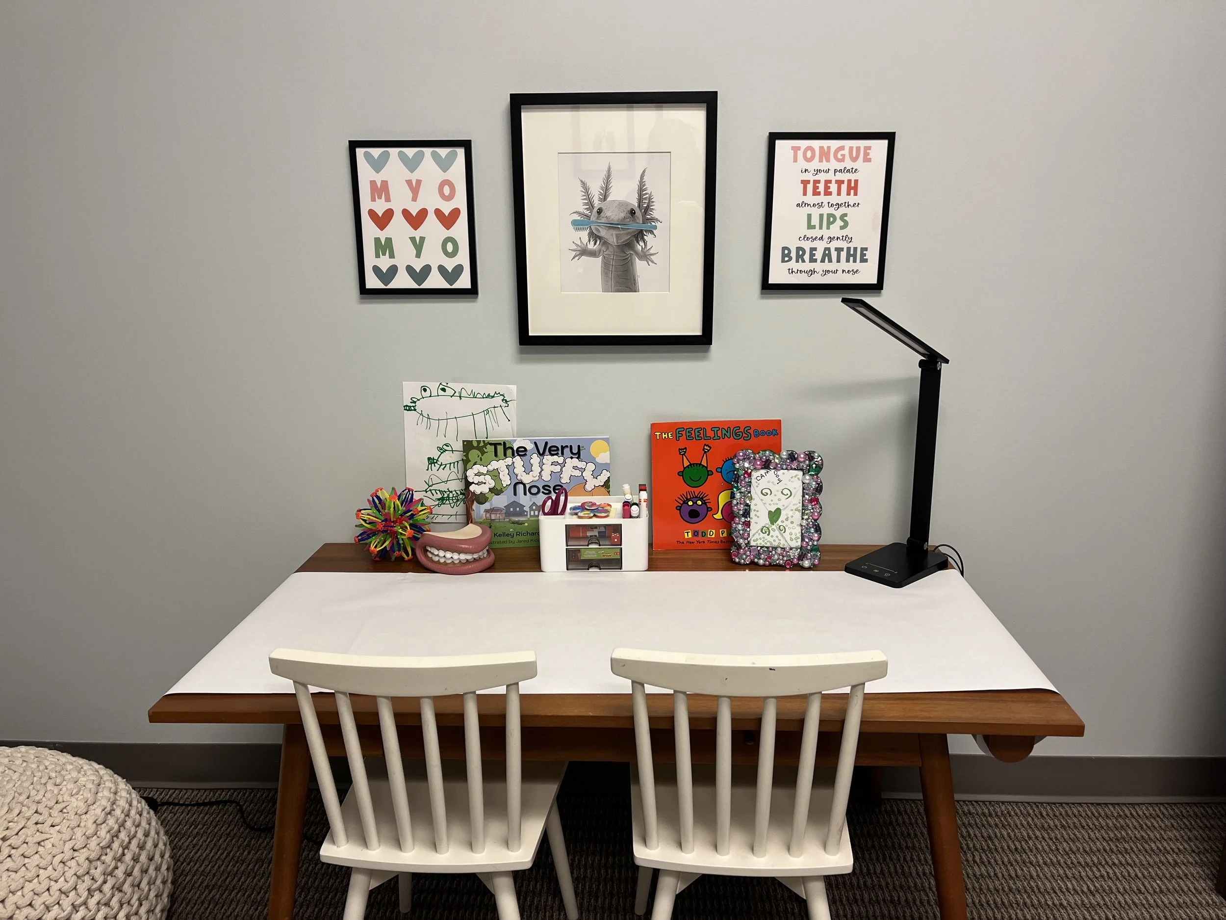 A children's craft and activity table with chairs, art supplies, books, and a desk lamp, decorated with wall art about emotions and the human body.