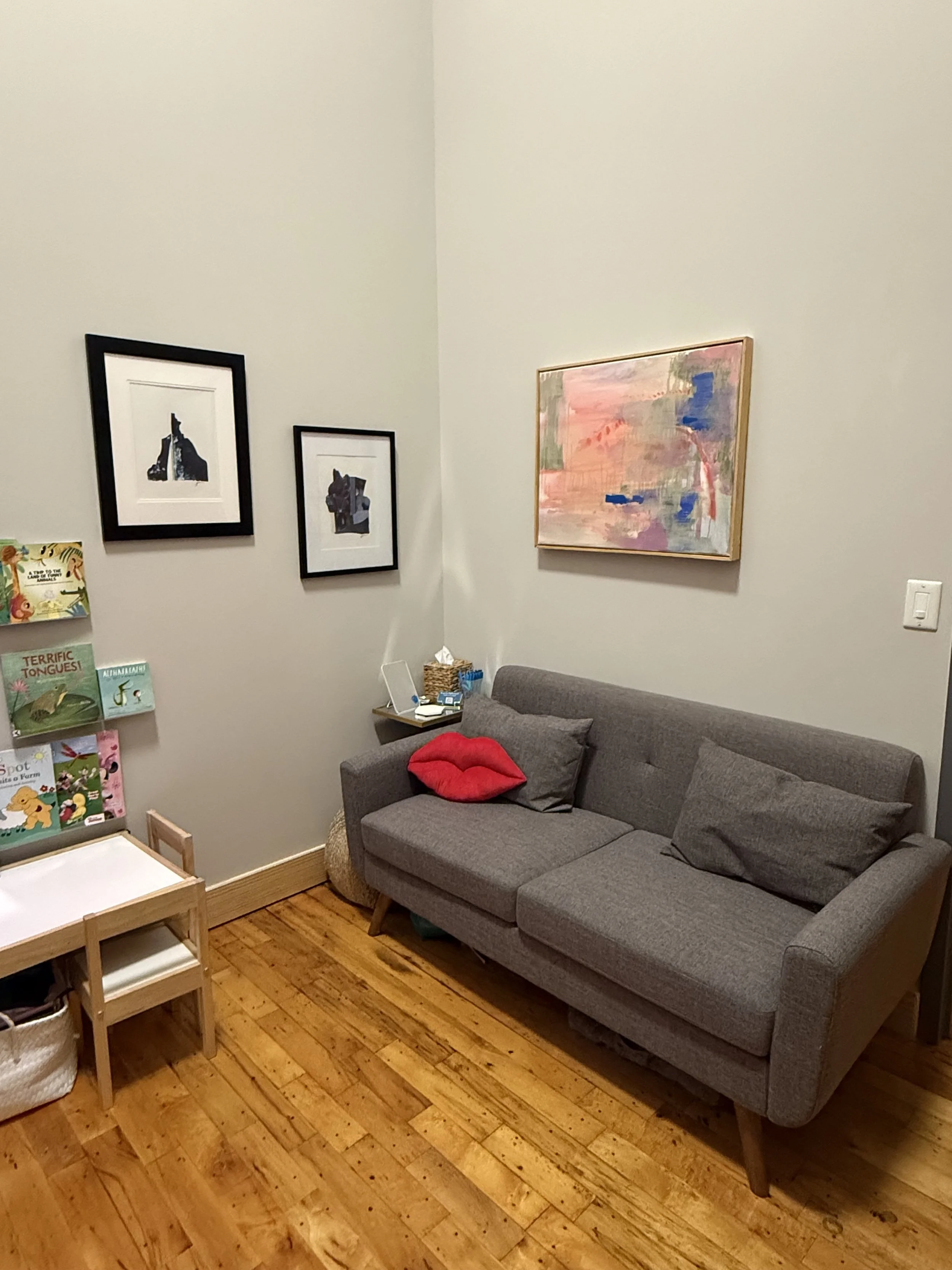 A cozy corner in a living room with a gray sofa, a cheeky lipstick-shaped pillow, and three throw pillows. There are four framed art pieces on the walls—two black and white abstract artworks and one pastel-colored abstract painting. A small shelf holds books and a wicker basket. The room has wooden flooring.