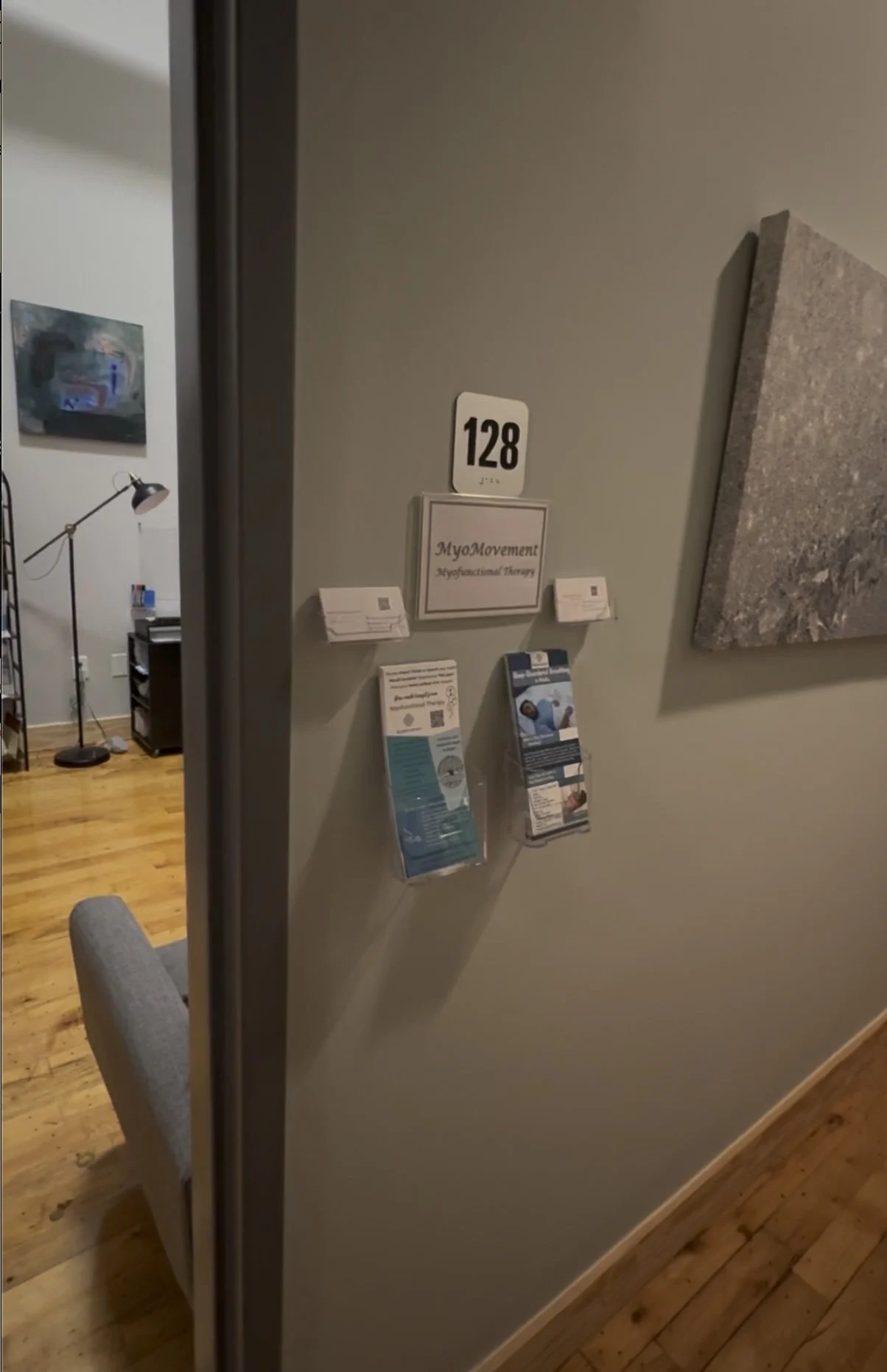 Indoor scene with wall displaying signage, brochures, and decorations. Part of a room with wooden floor, gray chair, and desk lamp visible.