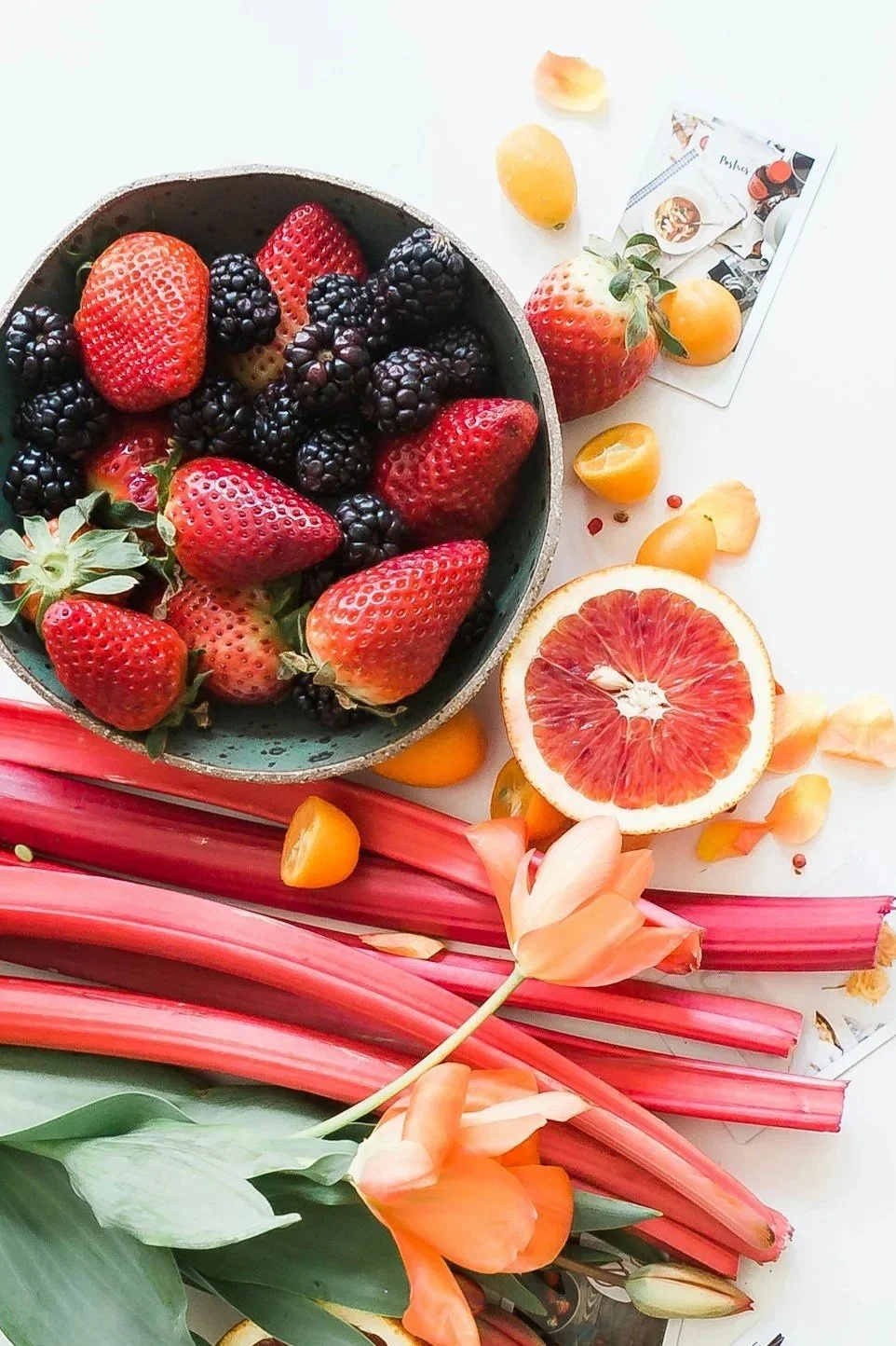 A bowl of mixed berries including strawberries, blackberries, and blueberries, along with a sliced grapefruit, surrounded by flowers, jewelry, and small decorative items on a white surface.
