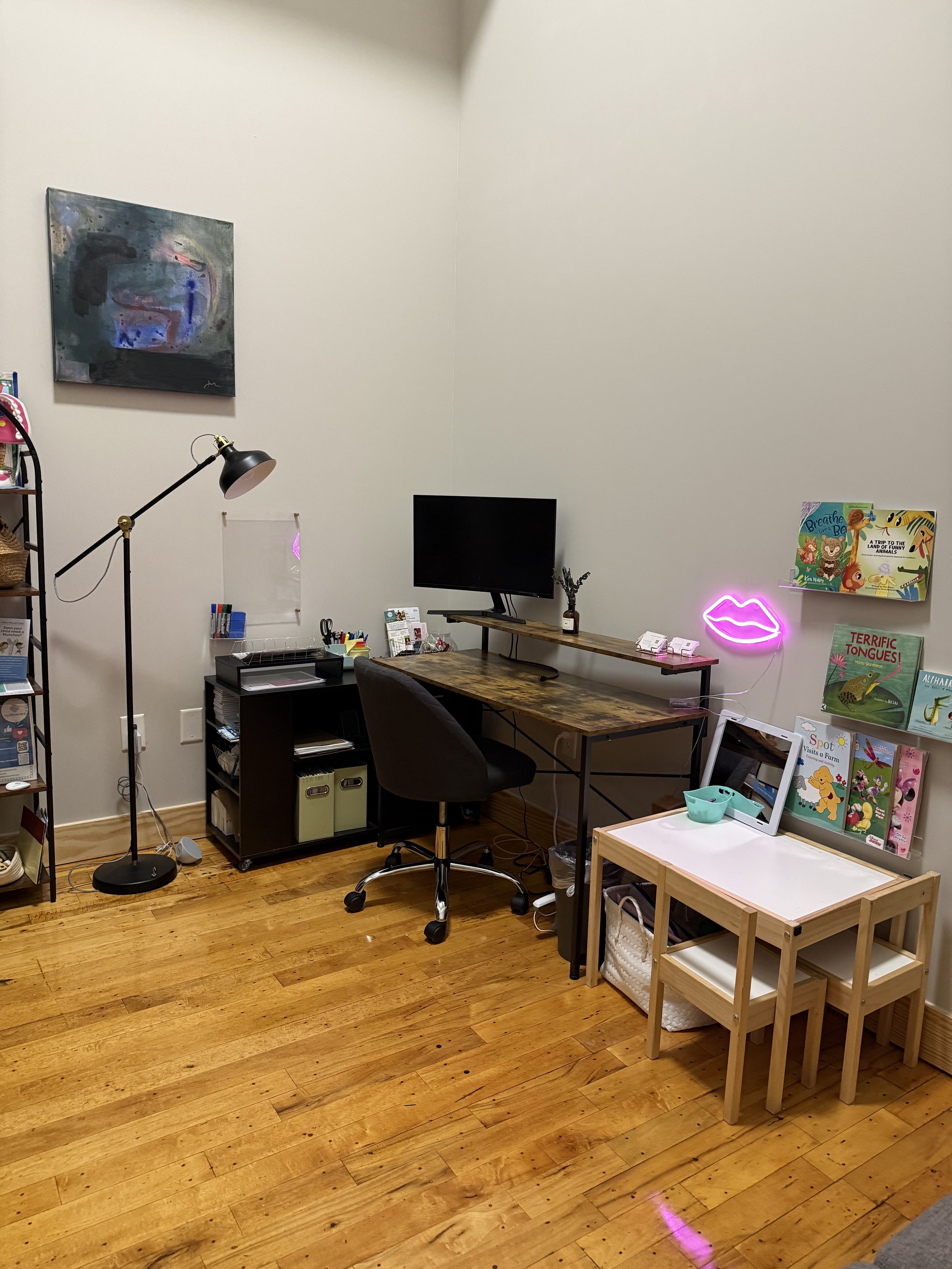 A cozy home office with a wooden desk, black office chair, small bookshelf, wall-mounted TV, neon pink lips sign, and children's books on a shelf, with hardwood floors and abstract art on the wall.