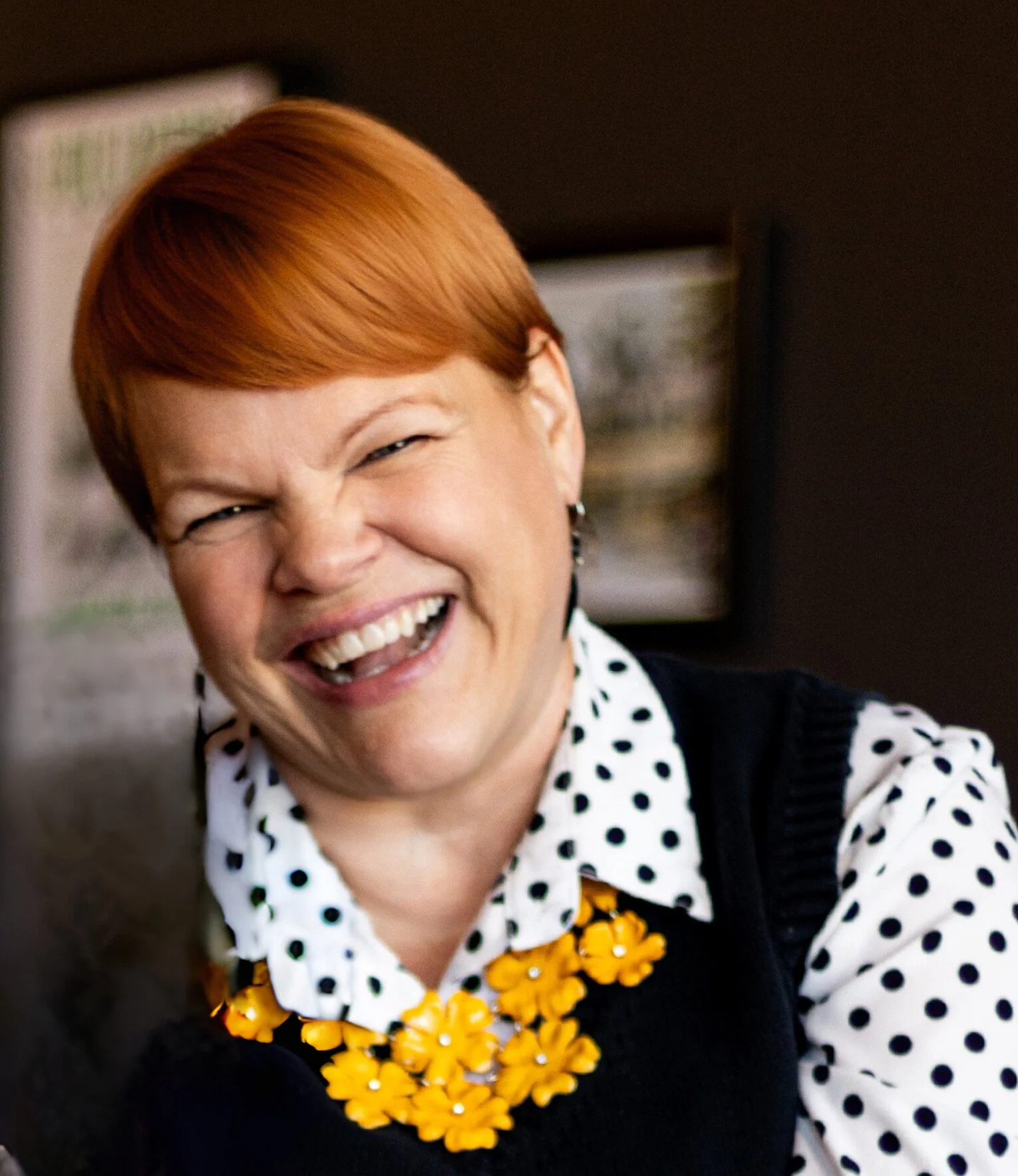 Close up of Jenna smiling, head titled, wearing a white shirt with black polkadots, a blacks sweater vest and yellow necklace.