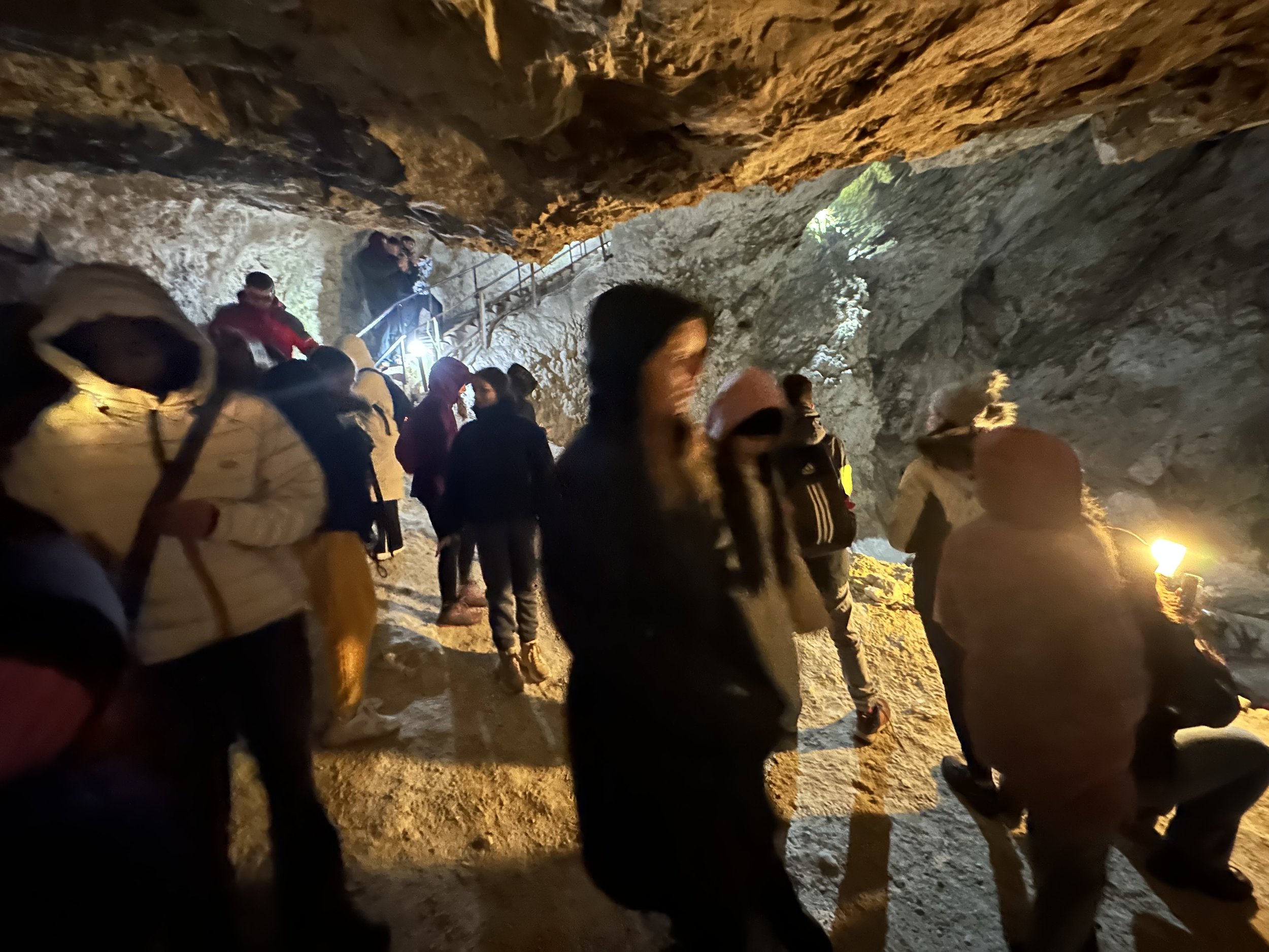 A group of people exploring a dimly lit cave with rough rocky walls, wearing winter clothing including jackets and hats. Some hold lanterns or flashlights to illuminate the path.