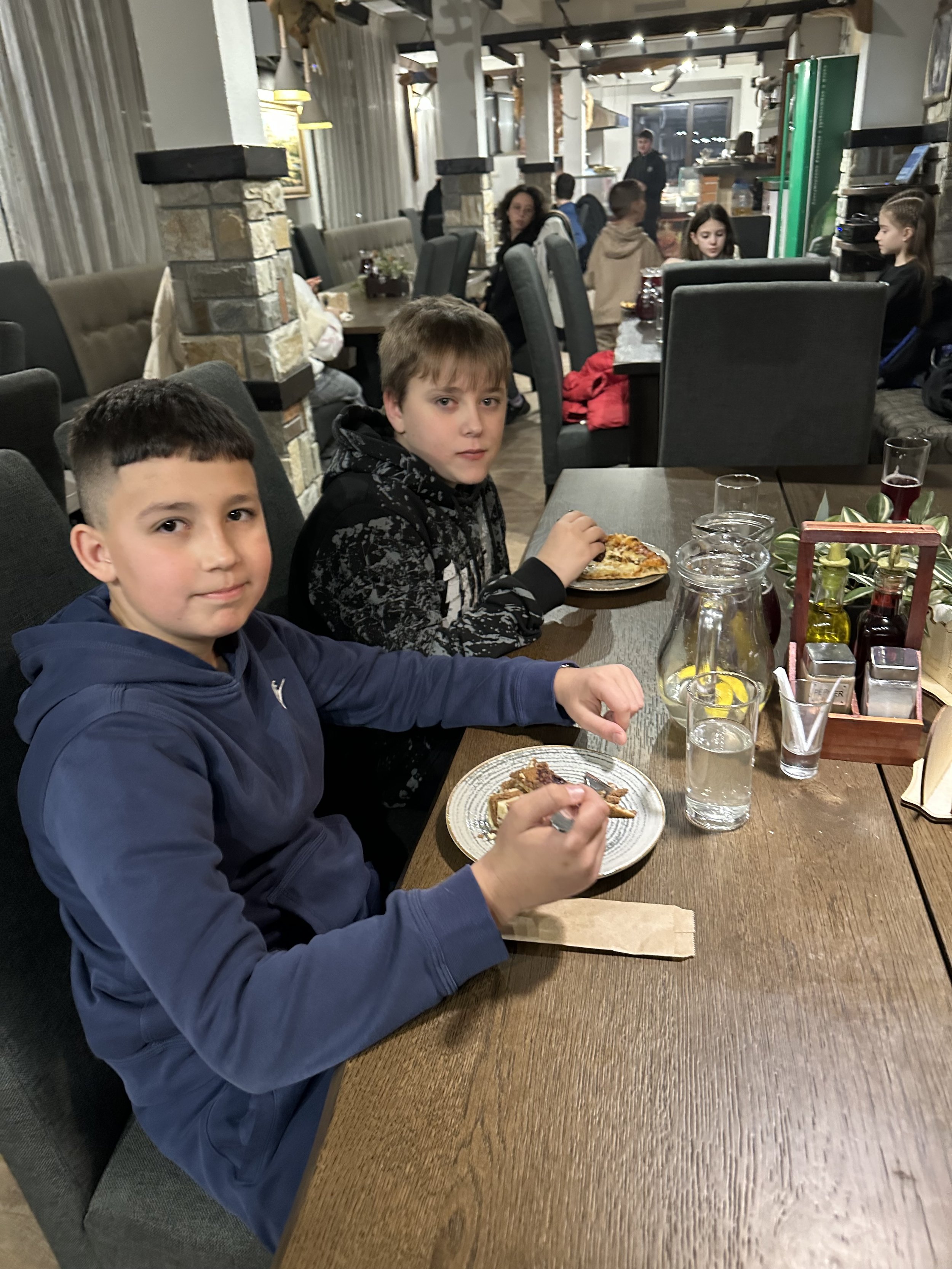 Two boys sitting at a restaurant table, eating a meal, with other people in the background.
