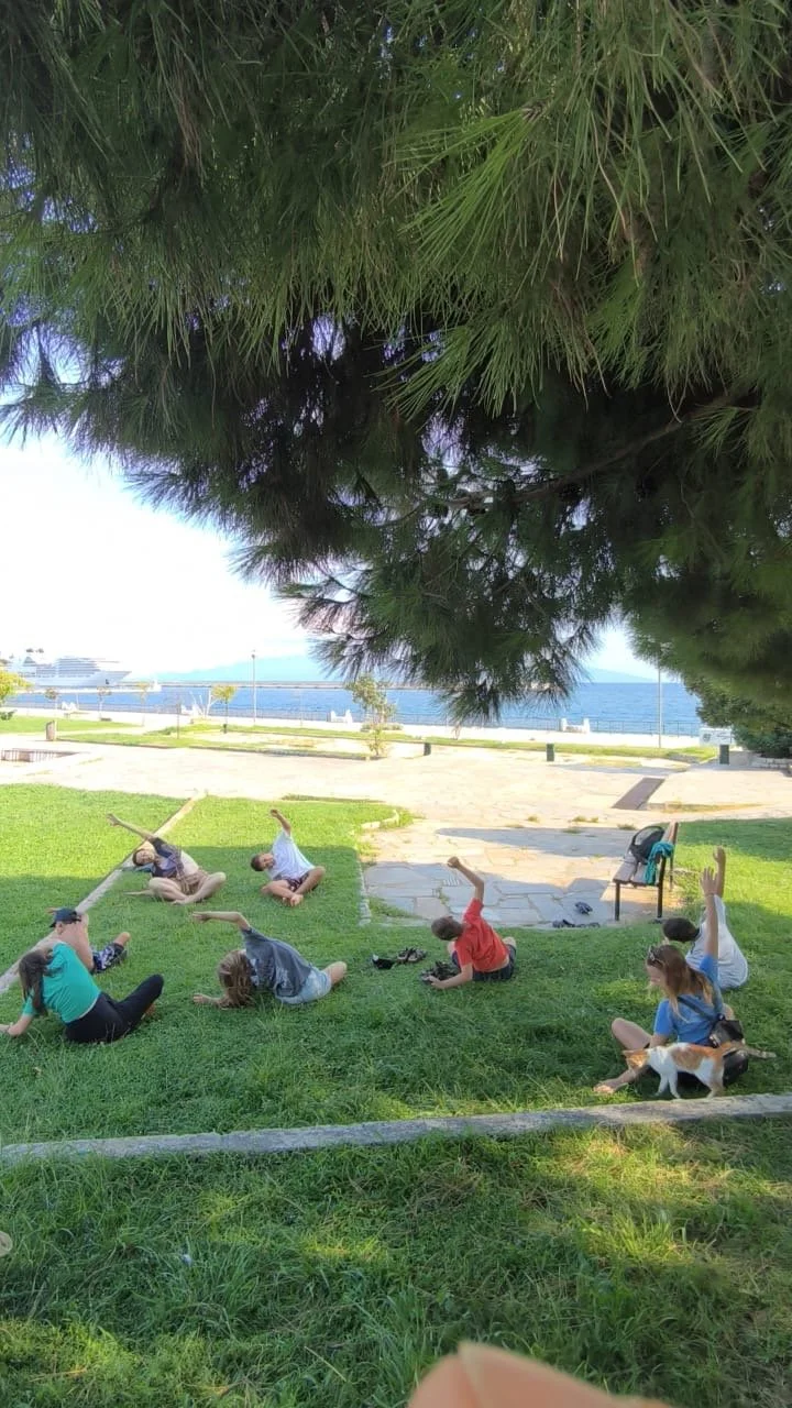 Children at Camp Evolve International practising yoga outdoors in Kavala, Greece, stretching and relaxing under a large tree by the water during a calm group activity.