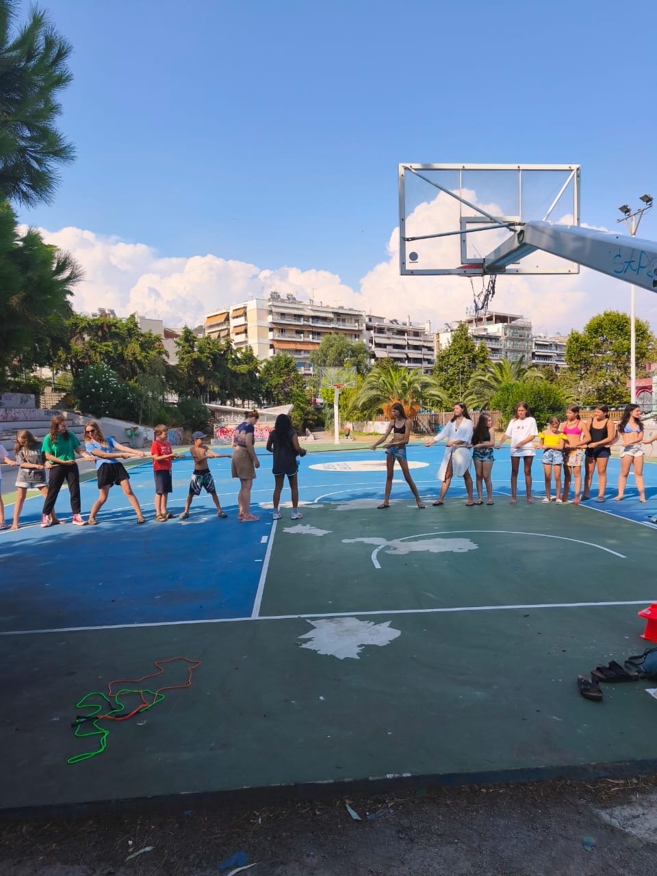 Children at Camp Evolve International taking part in a competition activity in Kavala, Greece, working together in a tug-of-war game on an outdoor court surrounded by trees and buildings under a bright blue sky.