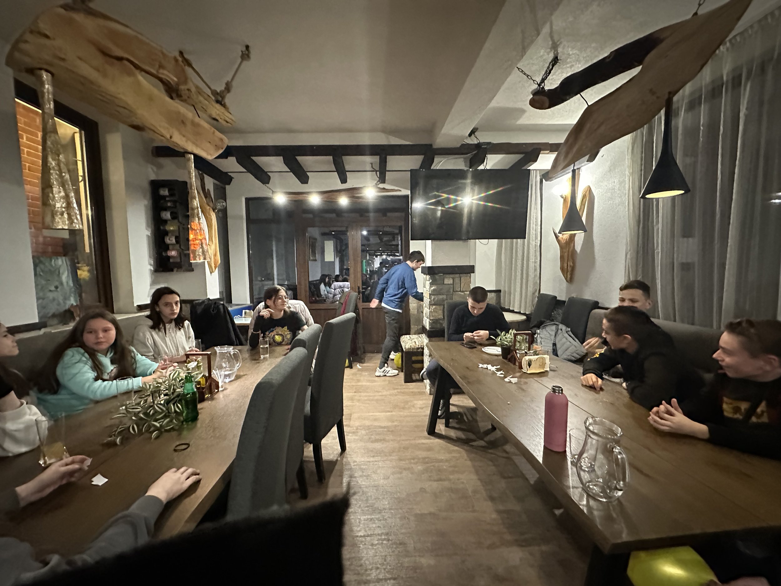 A group of people gathered around a long wooden table in a cozy indoor setting. The room features rustic wooden decor, pendant lighting, and a large TV mounted on the wall. Some people are engaged in conversation, while others are looking at their ph