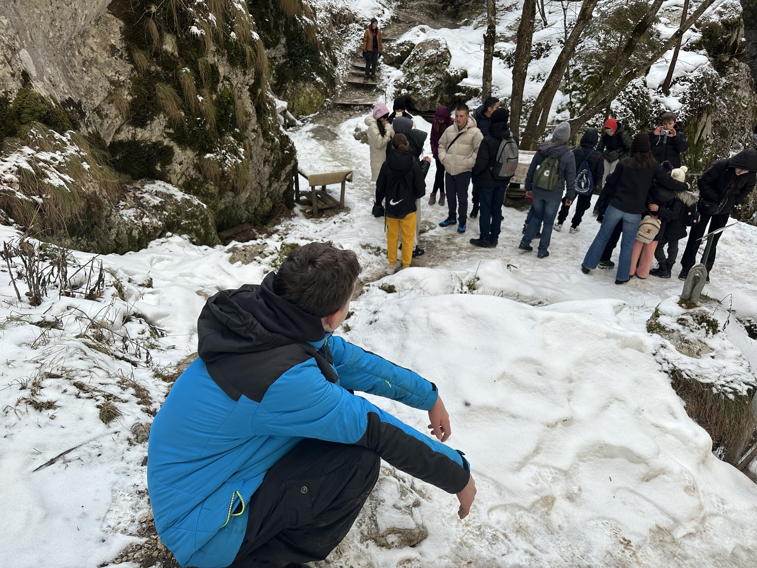 A group of people standing on a snowy path in a winter forest, with one person in a blue jacket crouching in the foreground.