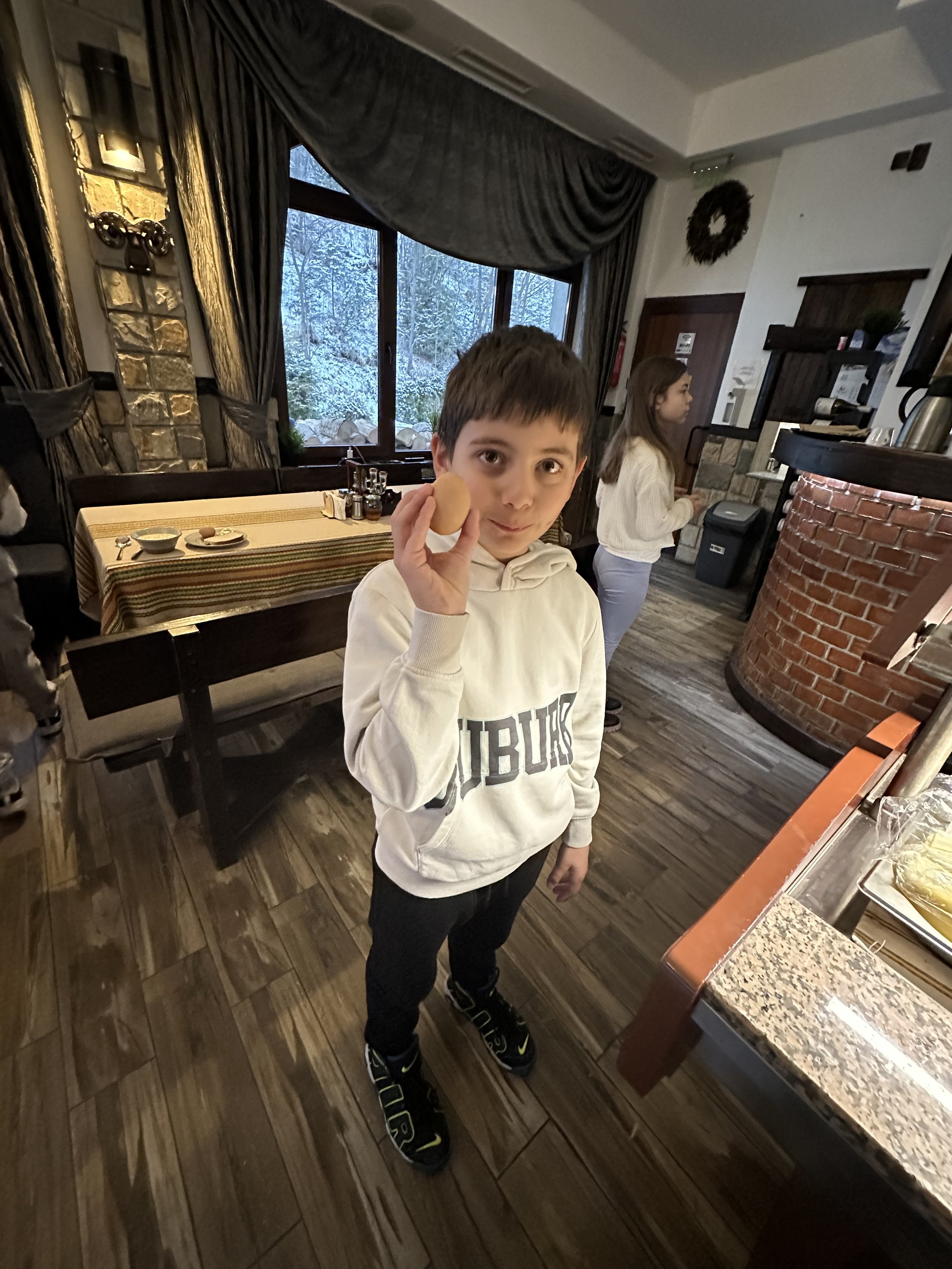 A young boy holding a brown egg in his hand while standing indoors in a room with wood flooring and a large window. Another person is visible in the background. The setting appears to be a restaurant or a cafΓ© with a table and chairs.