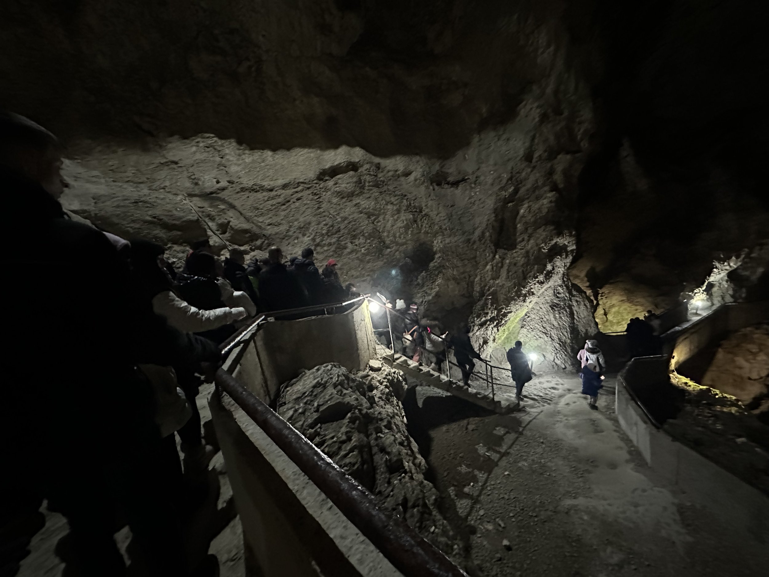 People exploring a dimly lit cave, walking along a pathway with handrails, surrounded by rock formations.