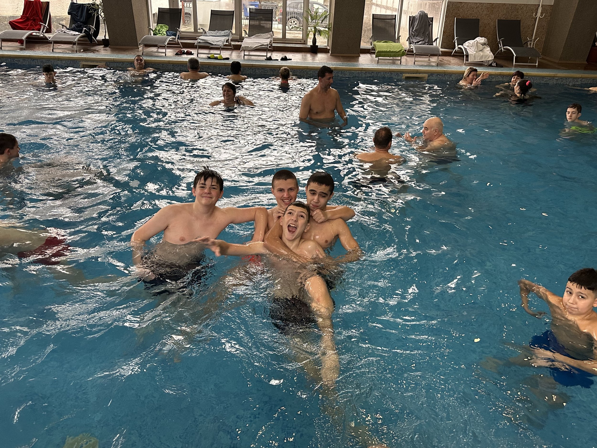 Group of people swimming in an indoor pool, with some lounging on chairs around the pool area.