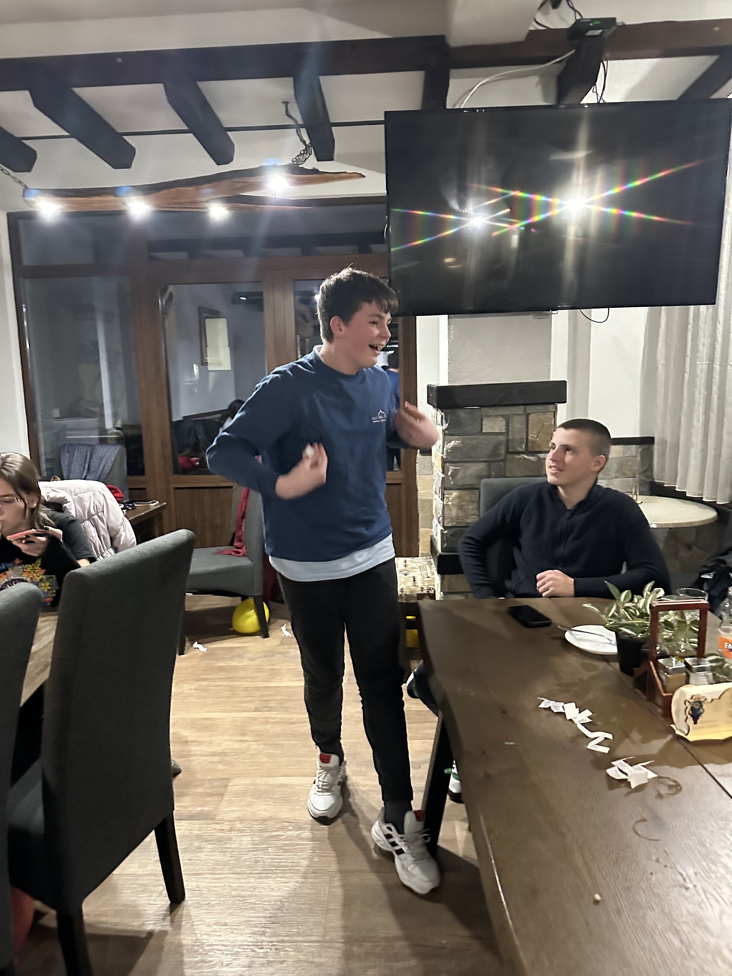 People gathered in a room, one standing and gesturing, others seated at a table with a TV and decor visible in the background.