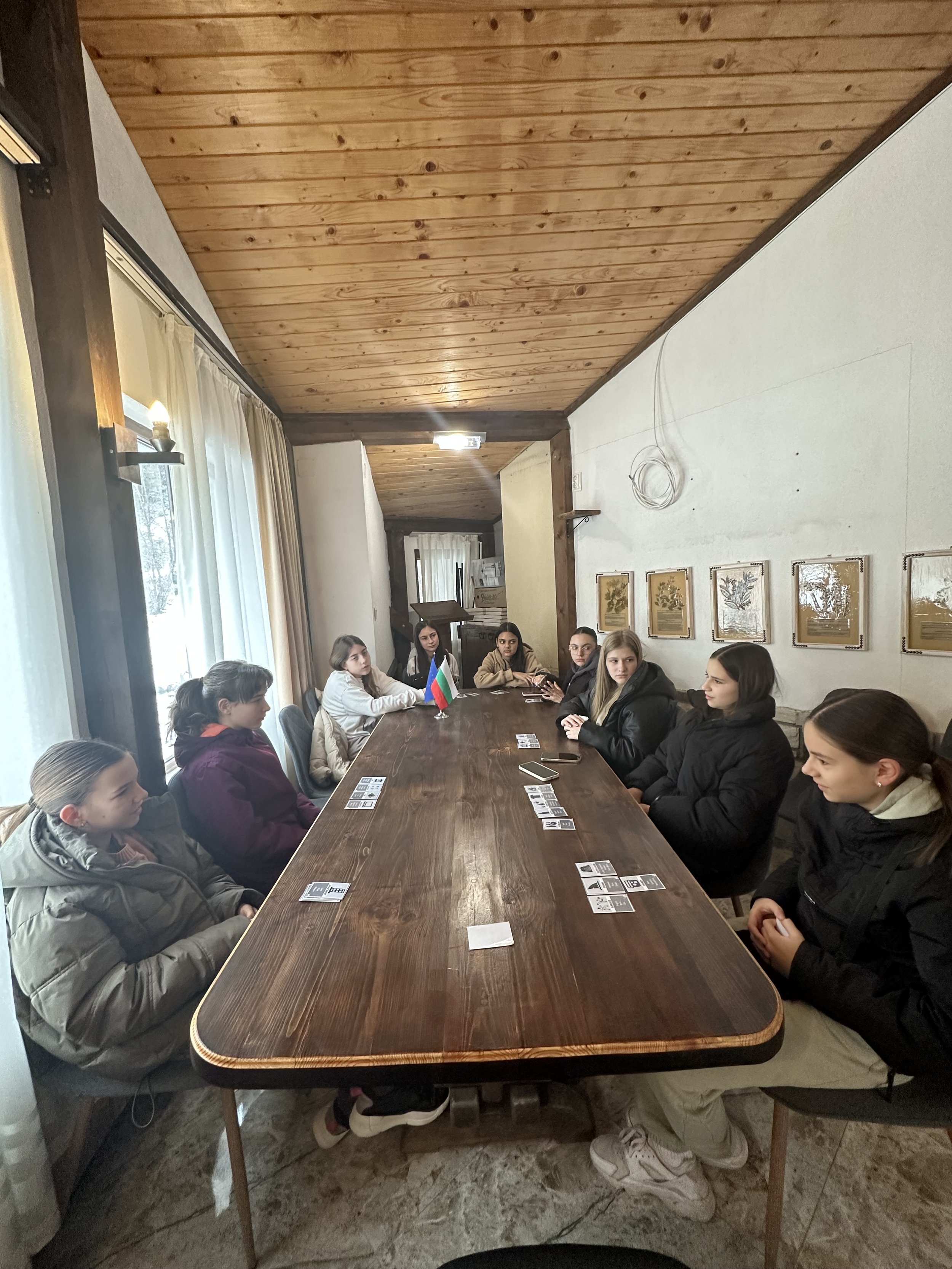 Group of people sitting around a long wooden table in a cozy room with a wooden ceiling, playing a card game. A small flag is on the table.