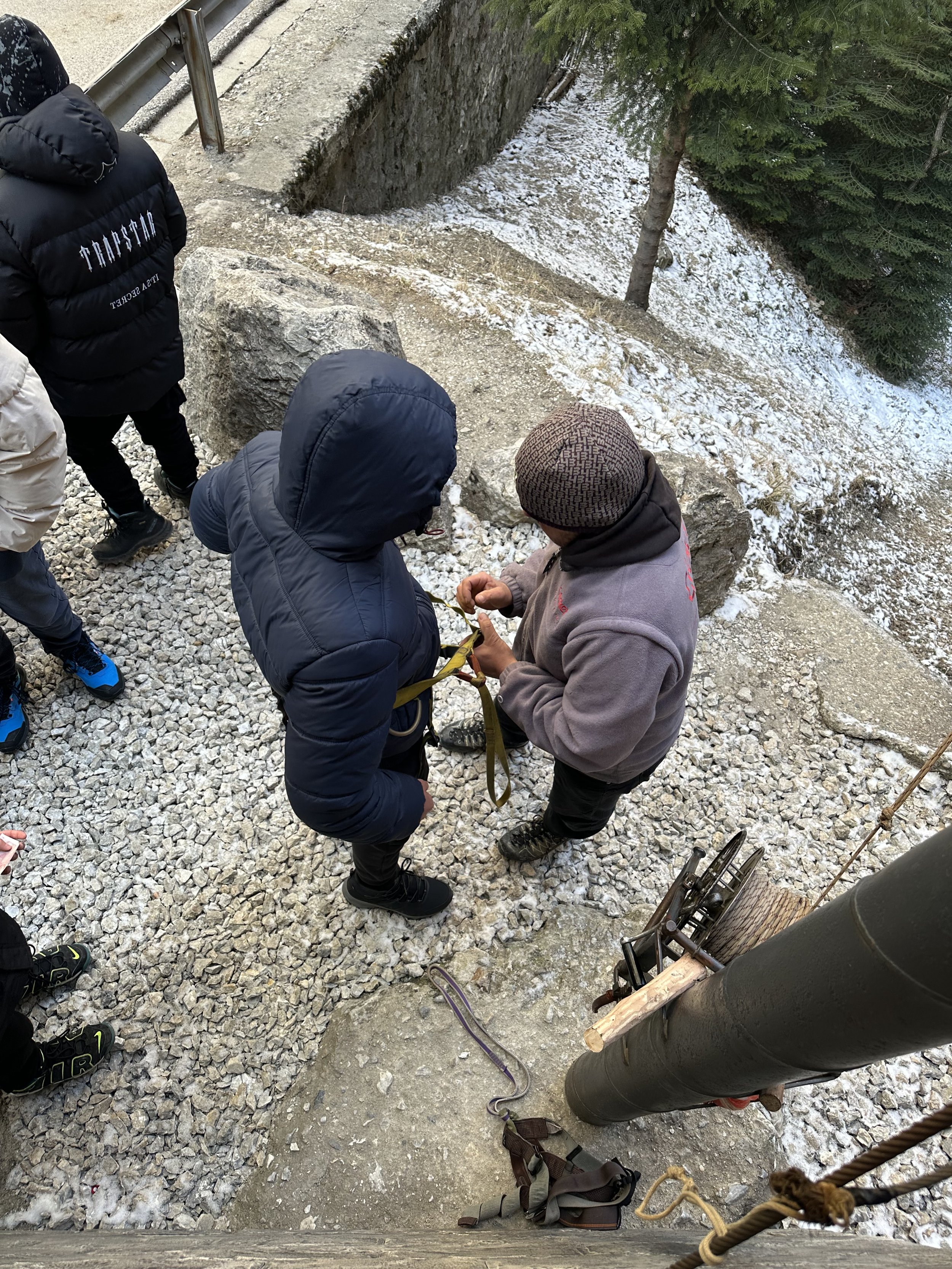 People near a rocky slope preparing climbing equipment, including harnesses and ropes, in a snowy outdoor setting, with winch apparatus visible.