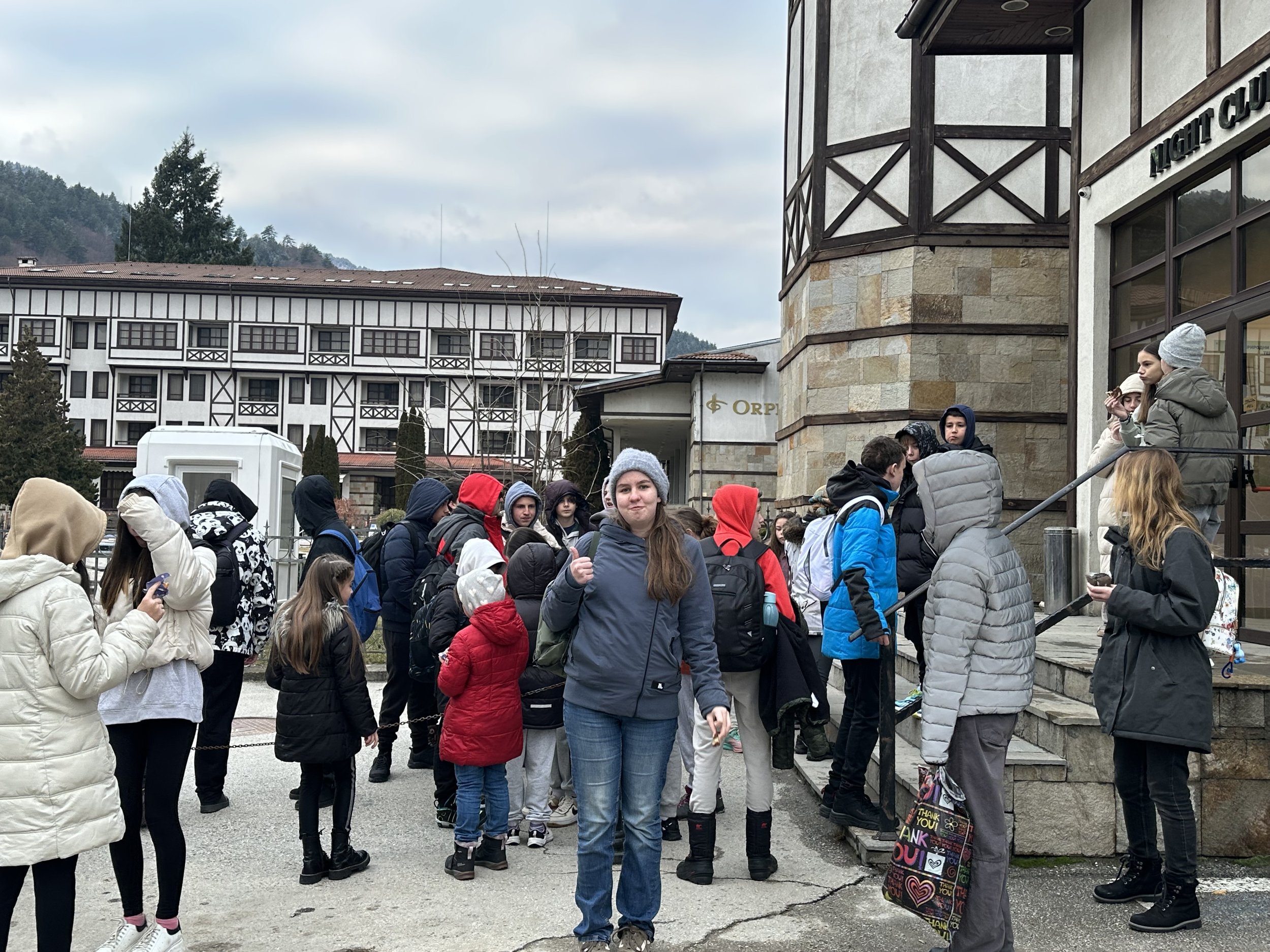 A large group of people, predominantly young, wearing winter jackets and hoodies, gathered outdoors near a stone building with a "Night Club" sign. The background shows traditional-style architecture and a partially cloudy sky.