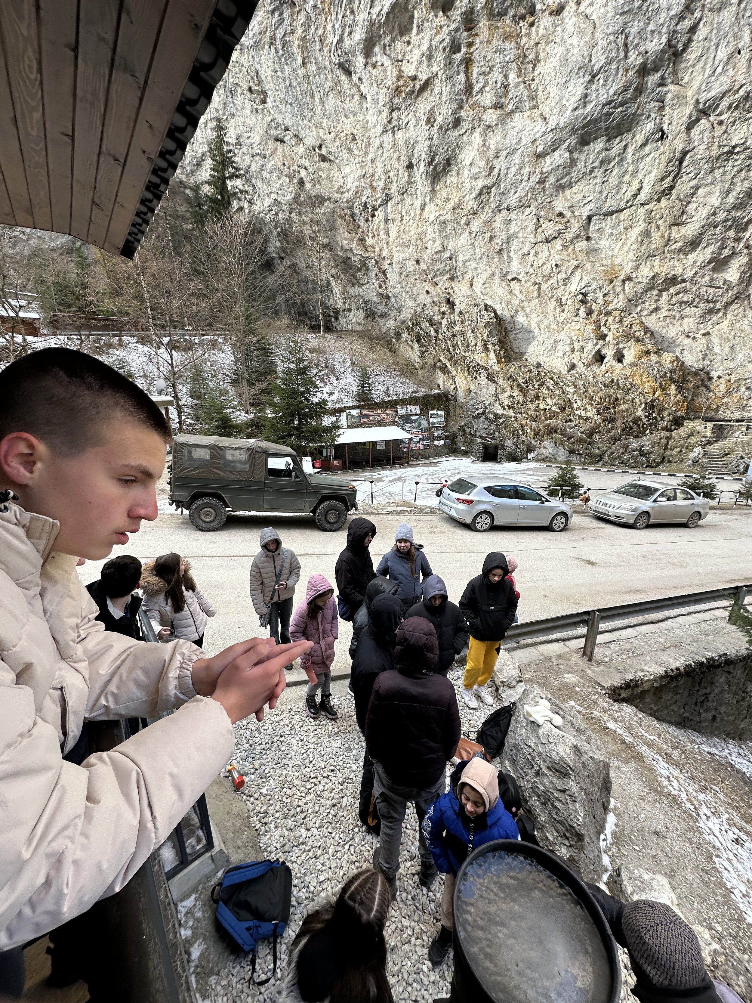 Group of people dressed in winter clothing gathered outside near rocky terrain and vehicles, with one person using a phone leaning on a ledge.