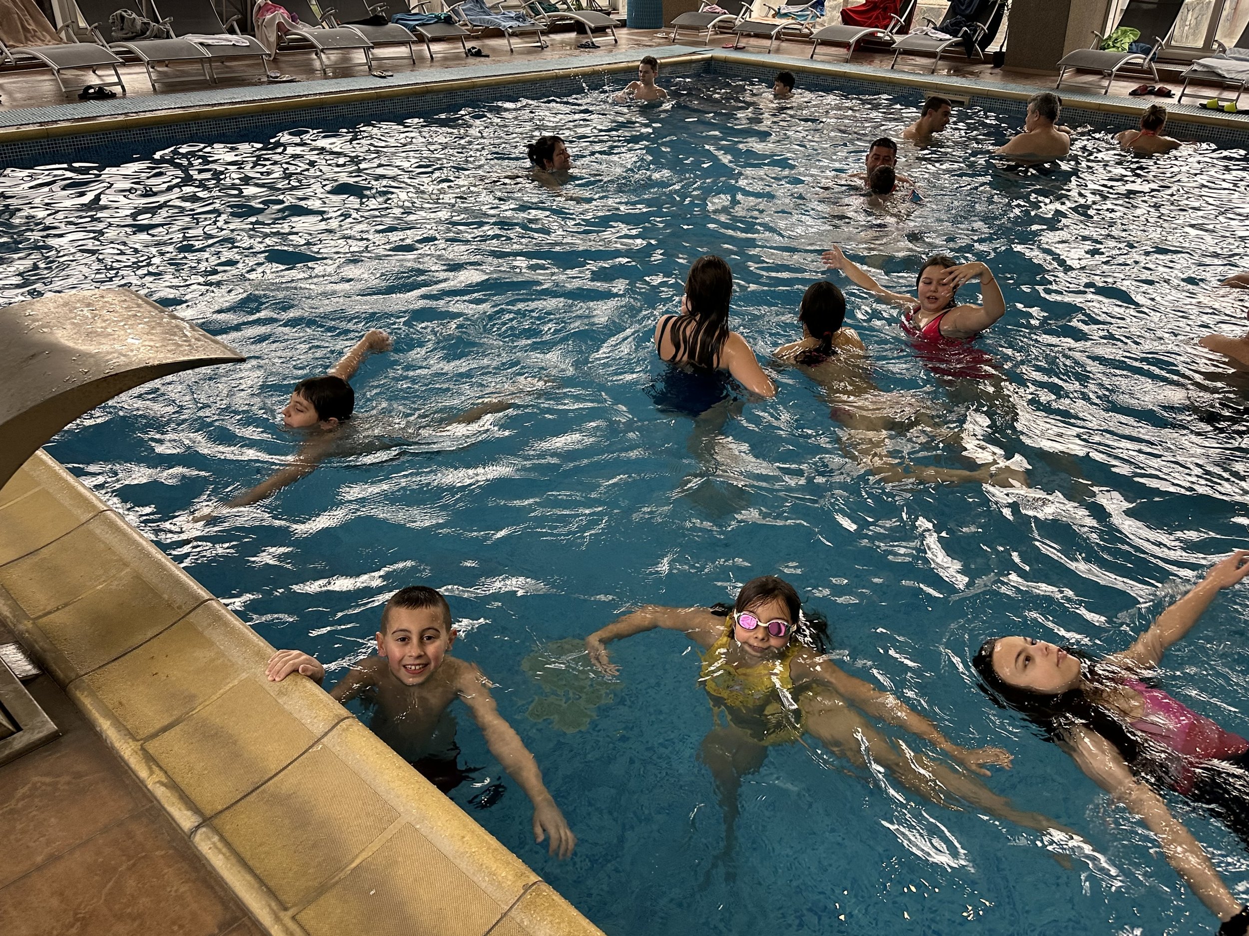 Indoor swimming pool with people swimming and relaxing, including children playing in the water. Poolside chairs visible in the background.
