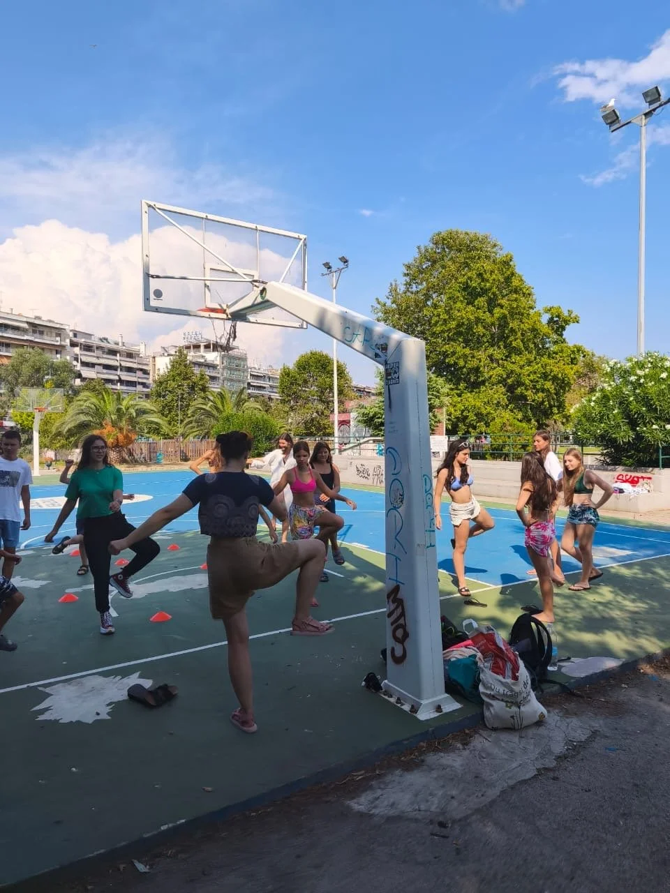 Children at Camp Evolve International taking part in an outdoor fitness activity in Kavala, Greece, exercising together on a basketball court under a clear blue sky surrounded by trees and buildings.