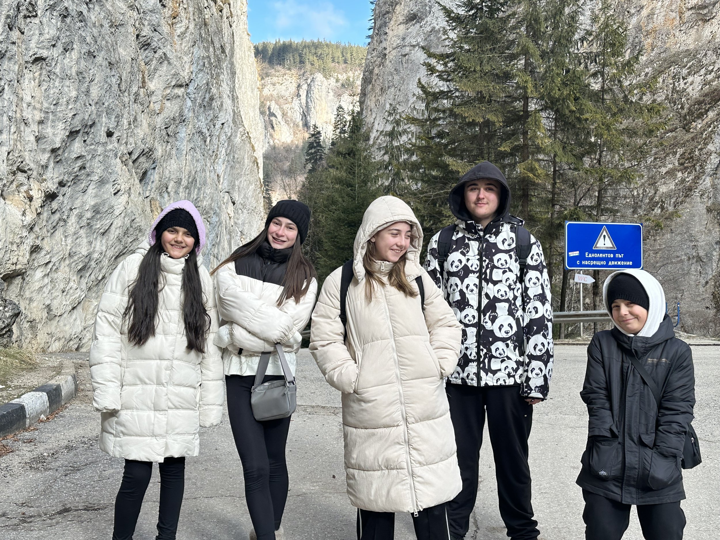 Group of people in winter clothing standing on a mountain road with rock cliffs and trees in the background.