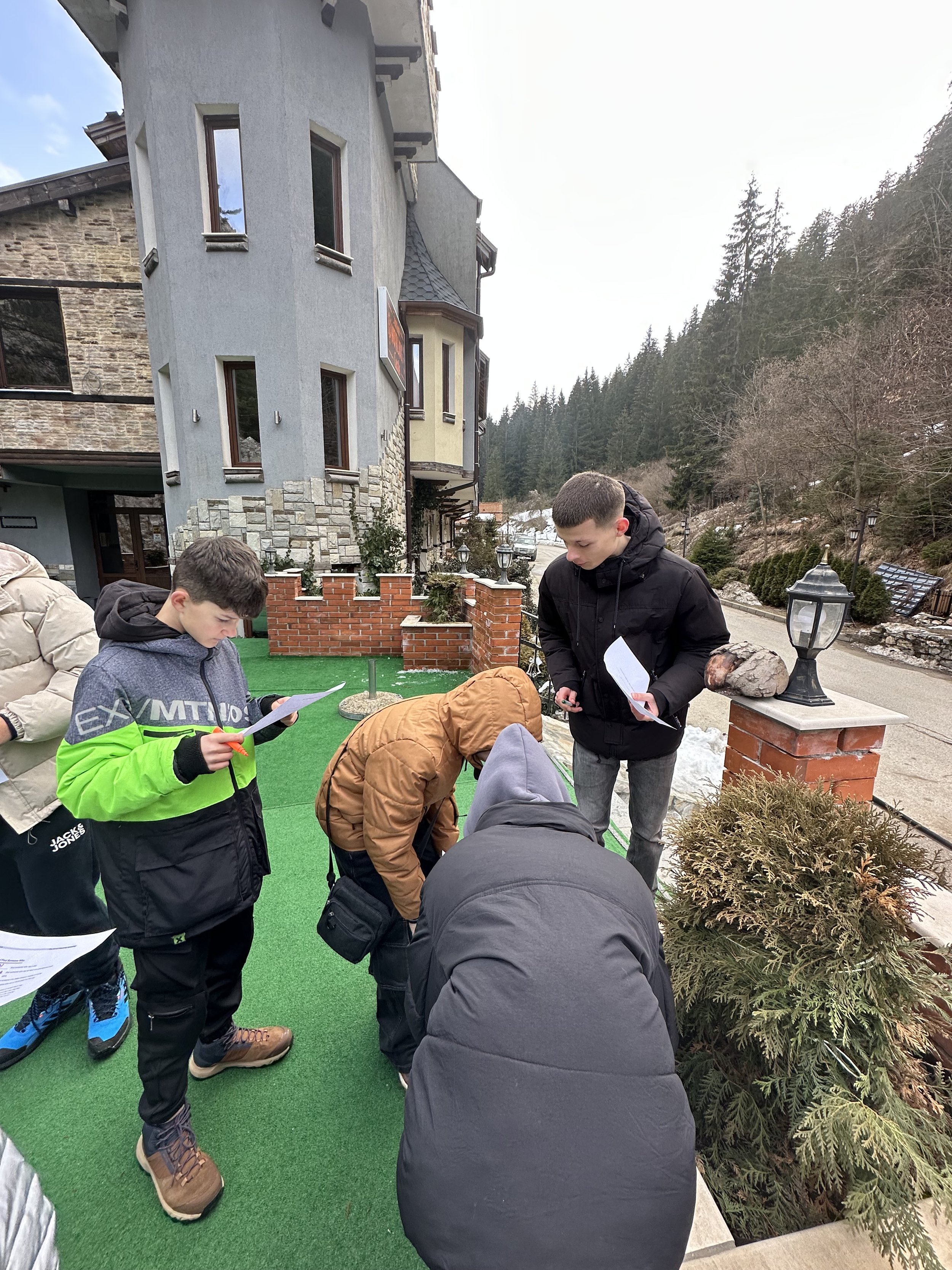 Group of people outdoors examining papers near a building and greenery.