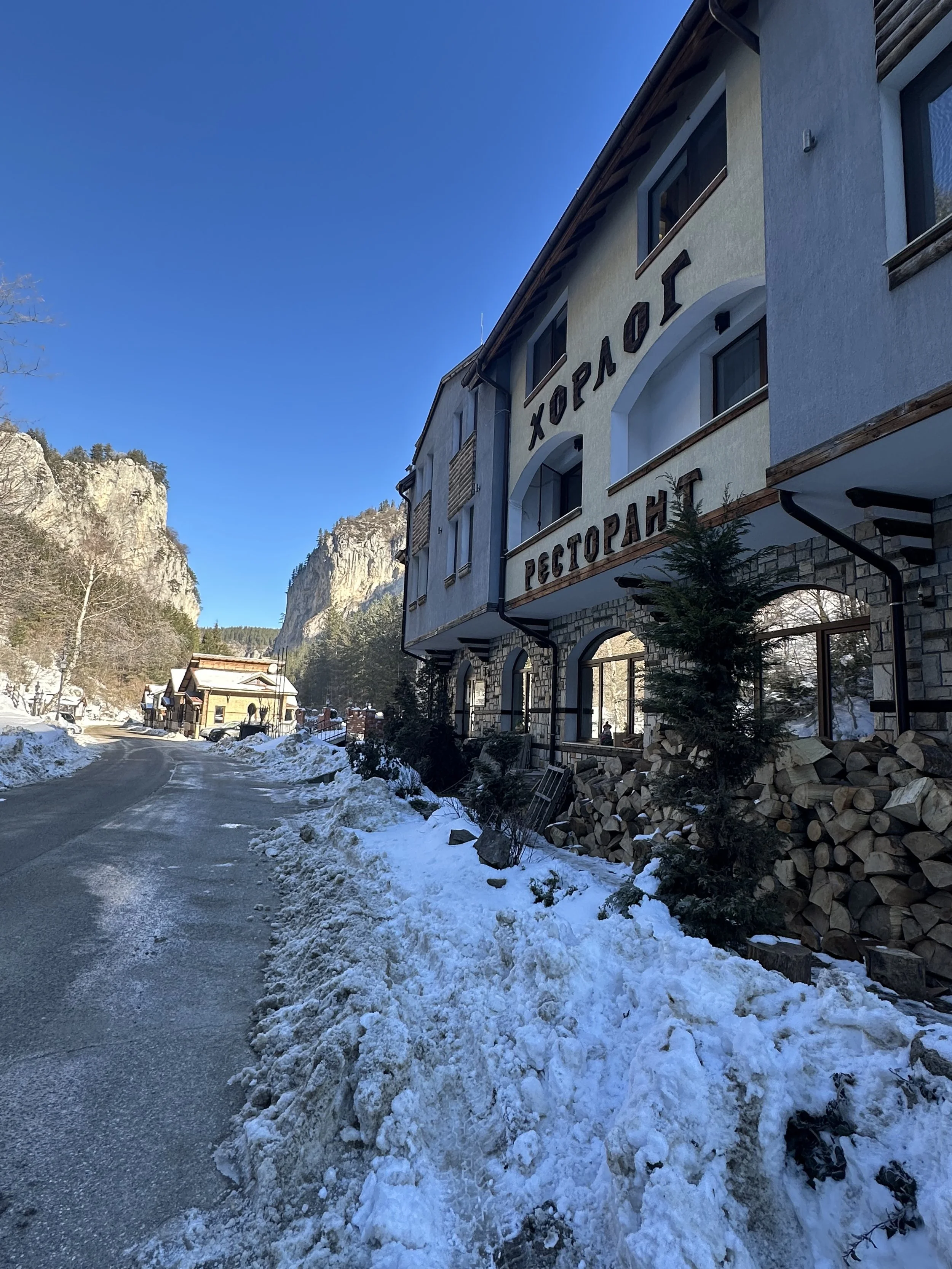 Snow-covered road leading past a building with a sign in Cyrillic, surrounded by mountains under a clear blue sky.