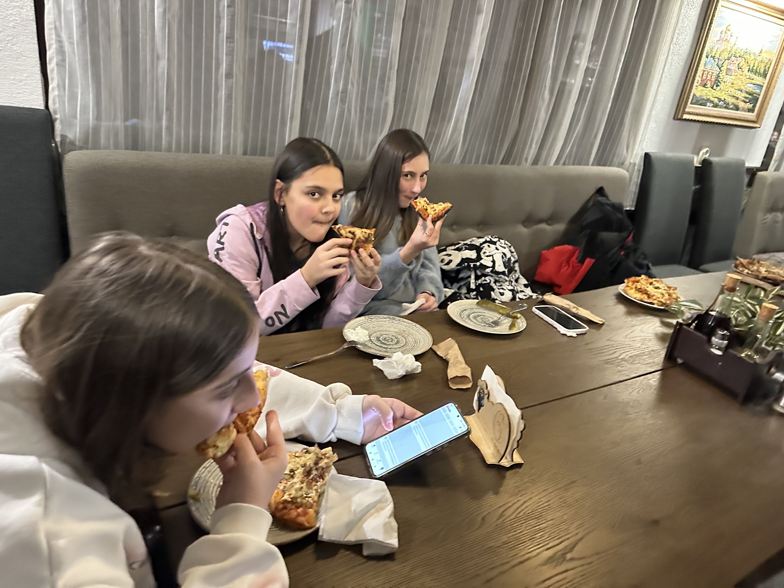 Three people eating pizza at a restaurant table, with phones and plates visible on the table.
