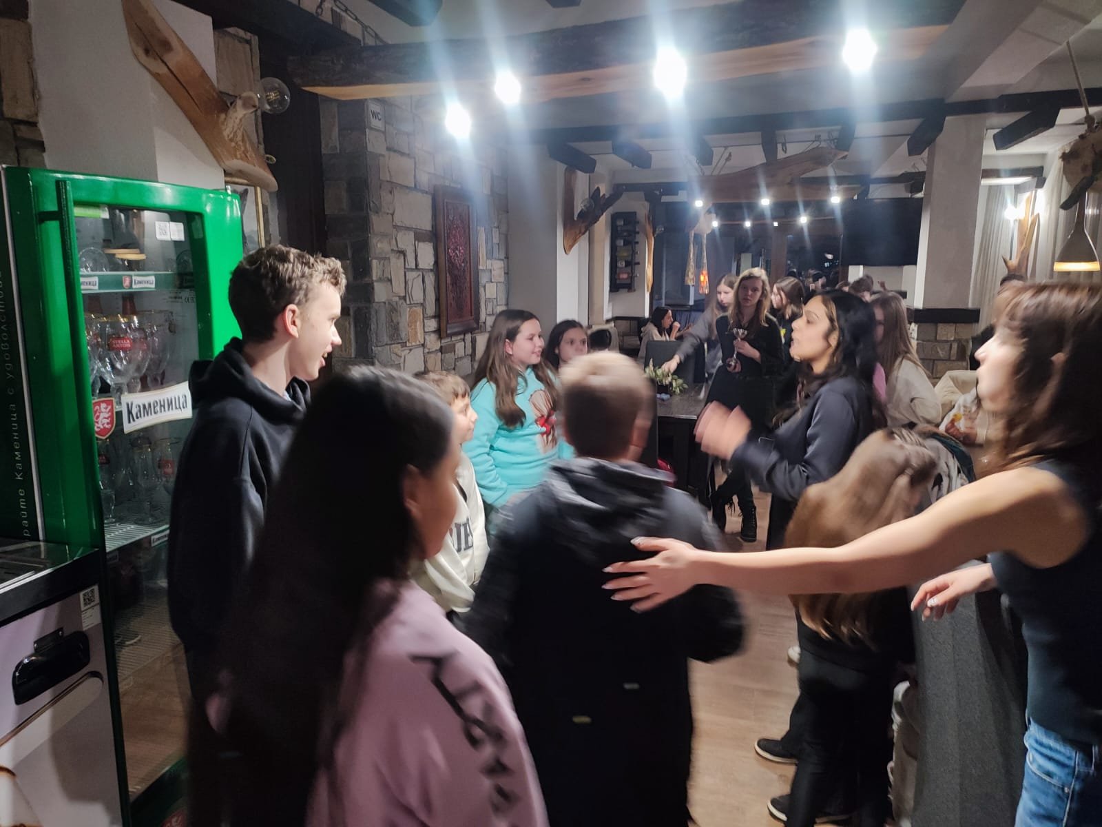 A group of people socializing indoors under bright lights in a rustic setting with wooden beams and stone walls. A green cooler labeled with a non-English brand is visible on the left. People of various ages, including children and adults, appear to