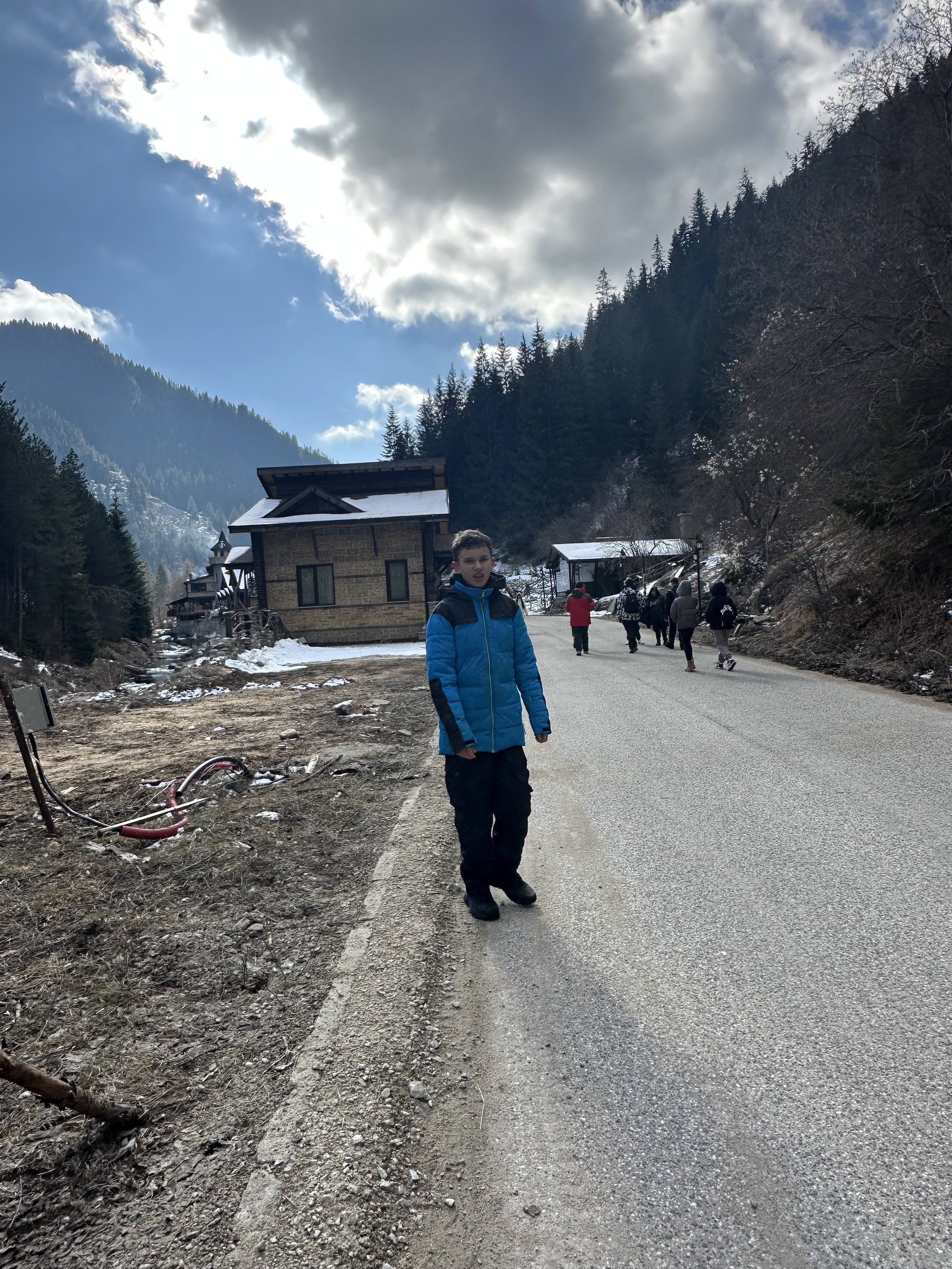 A person in a blue jacket standing on a road with wooden houses and pine trees in the background, under a partly cloudy sky.